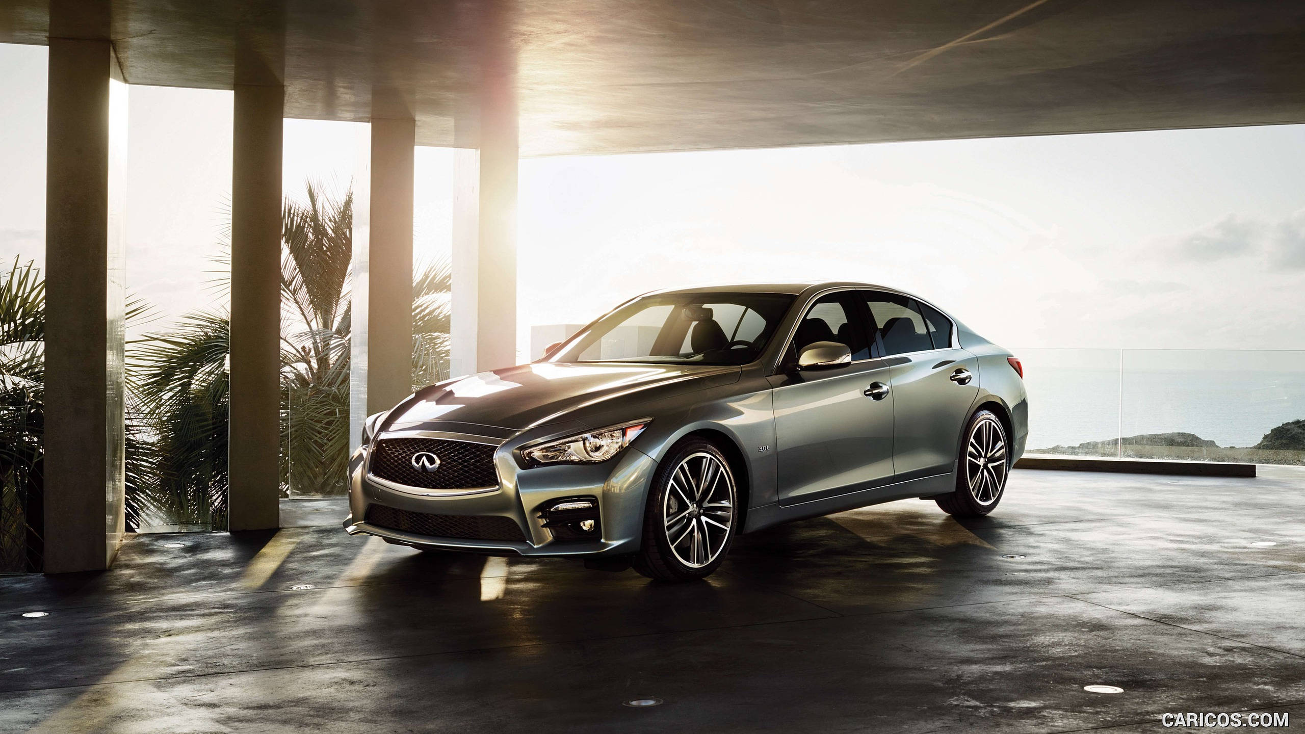 Infiniti 桌布 壁紙 — 一輛時尚的銀色轎車停放在現代混凝土結構下，周圍有棕櫚樹和海洋景觀，沐浴在金色陽光中。