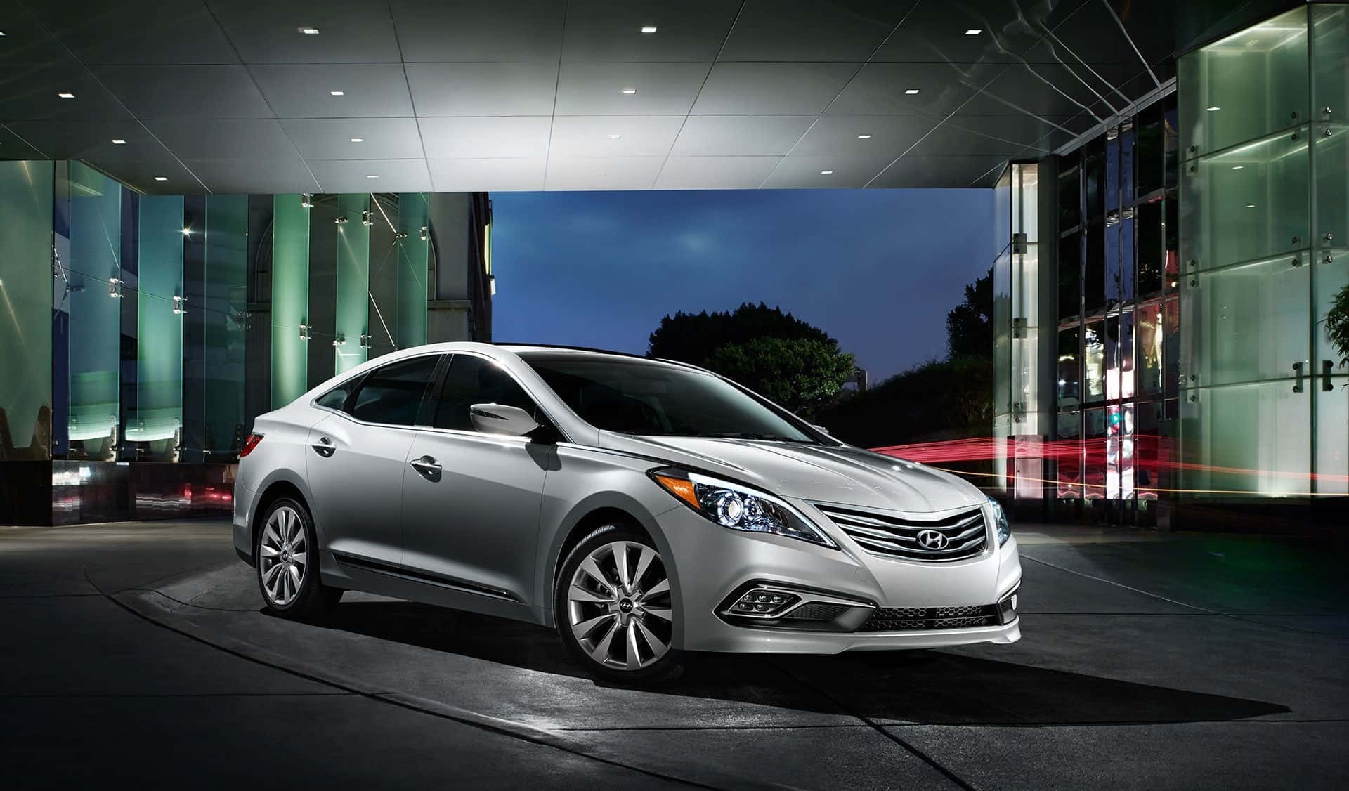 Hyundai Azera 桌布 壁紙 — 一輛時尚的銀色轎車在夜間燈光下閃耀，位於現代玻璃建築入口，帶有動態模糊條紋。
