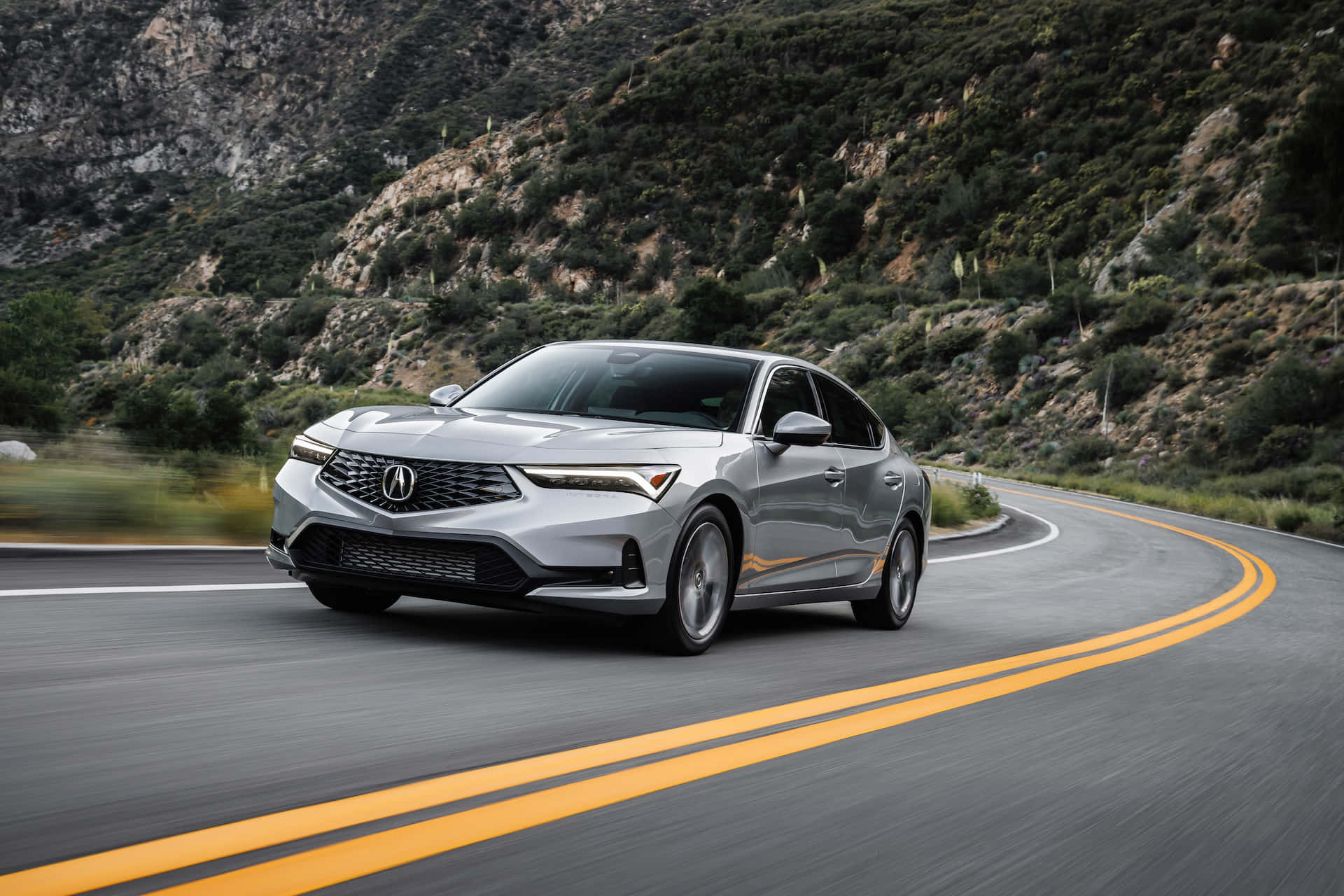 Acura Integra 桌布 壁紙 — 一輛時尚的銀色轎車在蜿蜒的山路上行駛，背景是模糊的綠樹和岩石地形。