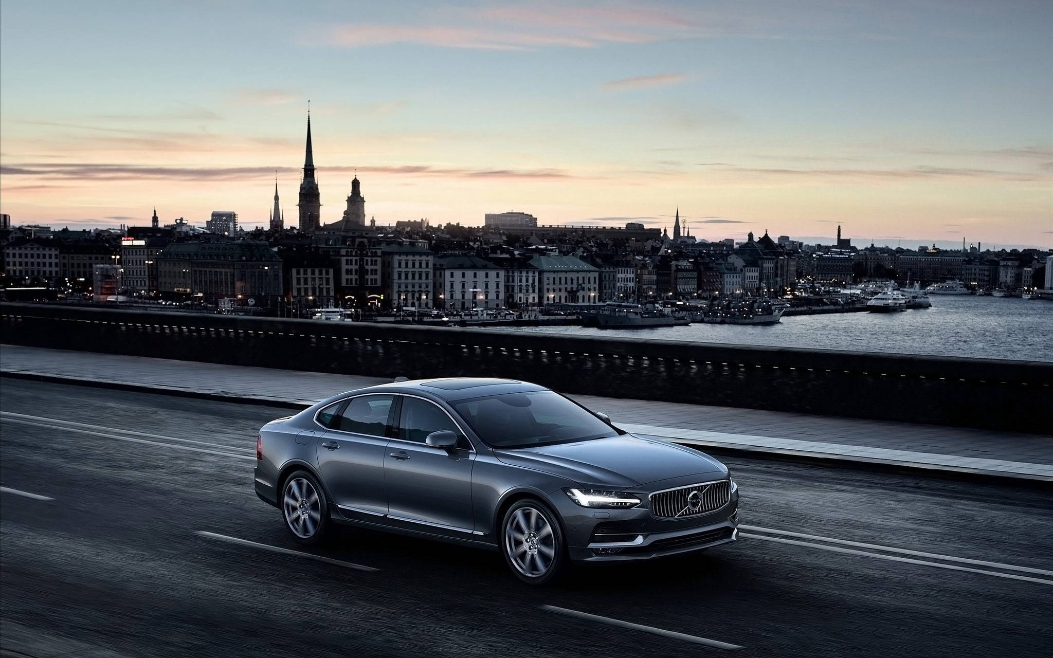 Volvo 桌布 壁紙 — 一輛時尚的銀色轎車沿著海濱道路行駛，城市天際線在夕陽下閃耀。
