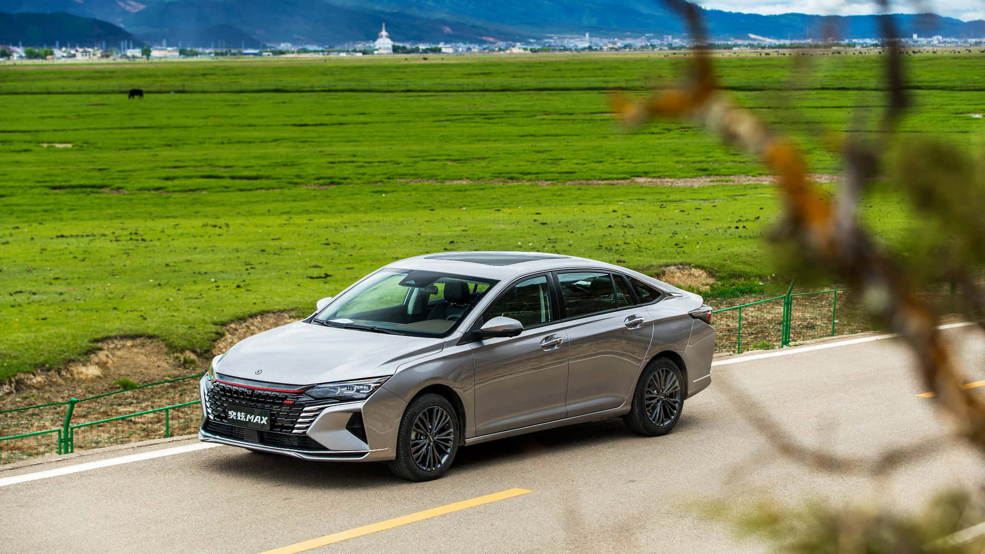 Dongfeng 桌布 壁紙 — 一輛時尚的銀色轎車沿著蜿蜒的道路行駛，穿過廣闊的綠色田野，遠處有山脈和一座白色寺廟，天空明亮。