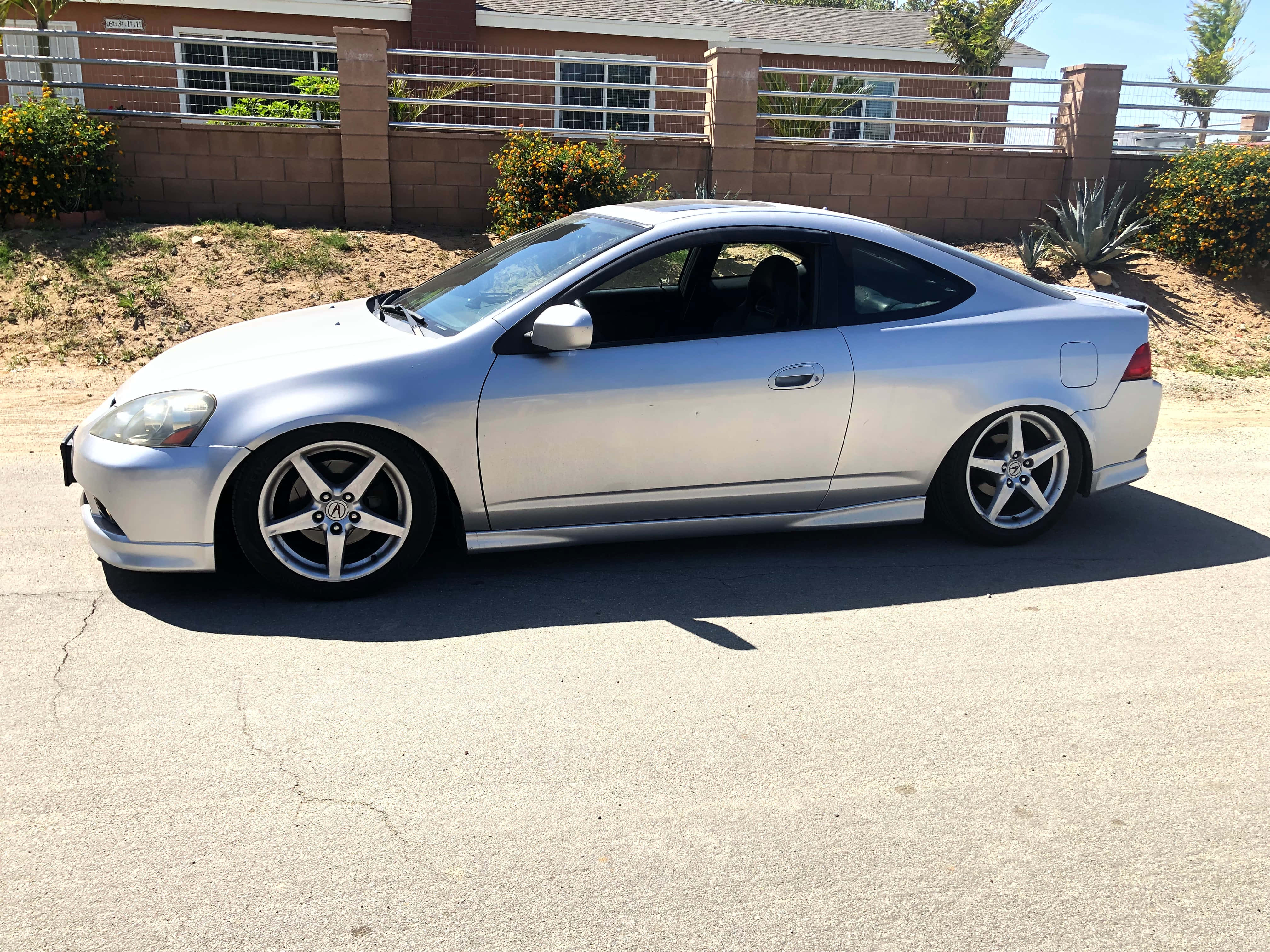 Acura Rsx 桌布 壁紙 — 一輛時尚的銀色運動轎跑車停在裂開的路面上，陽光明媚，背景有住宅圍欄和沙漠植物。