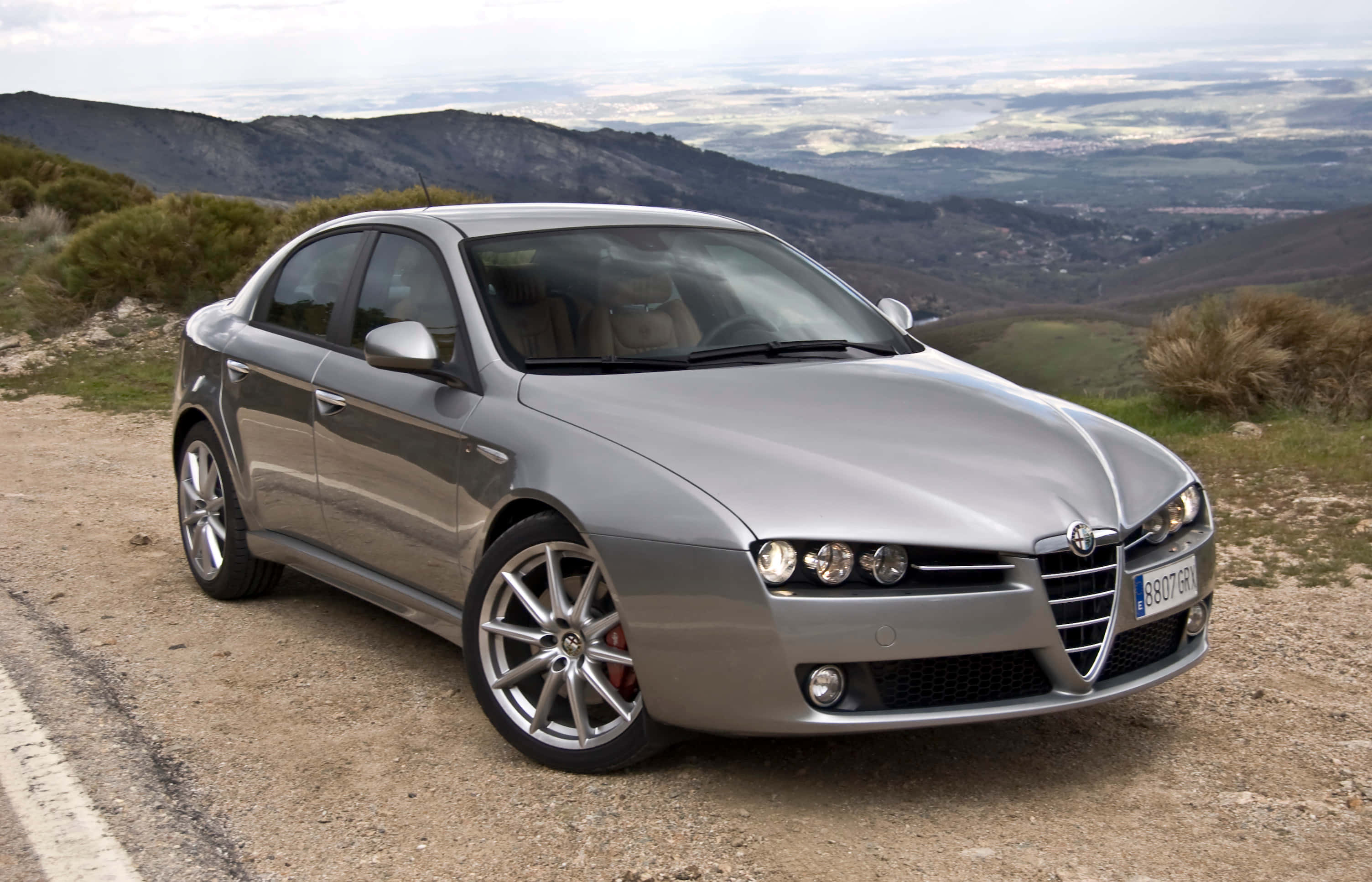 Alfa Romeo 159 桌布 壁紙 — 一輛時尚的銀色阿爾法羅密歐轎車停在一條土路上，俯瞰著多雲天空下的連綿起伏的丘陵。