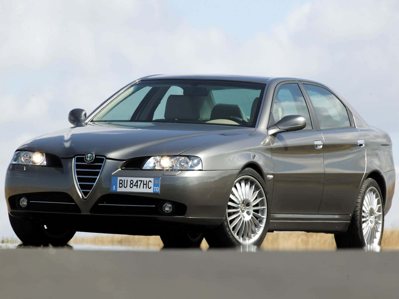 Alfa Romeo 156 桌布 壁紙 — 一輛時尚的銀色阿爾法羅密歐轎車在明亮的日光下閃耀，車頭燈亮起，映襯著蒼白的天空和乾草。