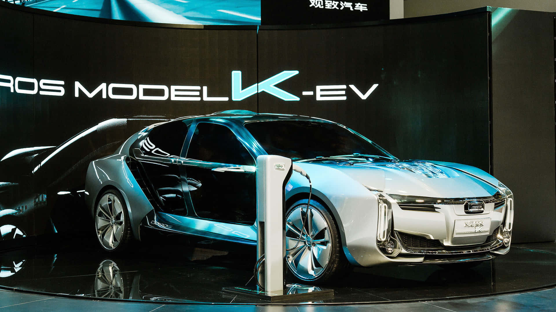Qoros 桌布 壁紙 — 一輛時尚的銀色電動車帶有發光藍色裝飾，正在高科技車展上連接到充電站。