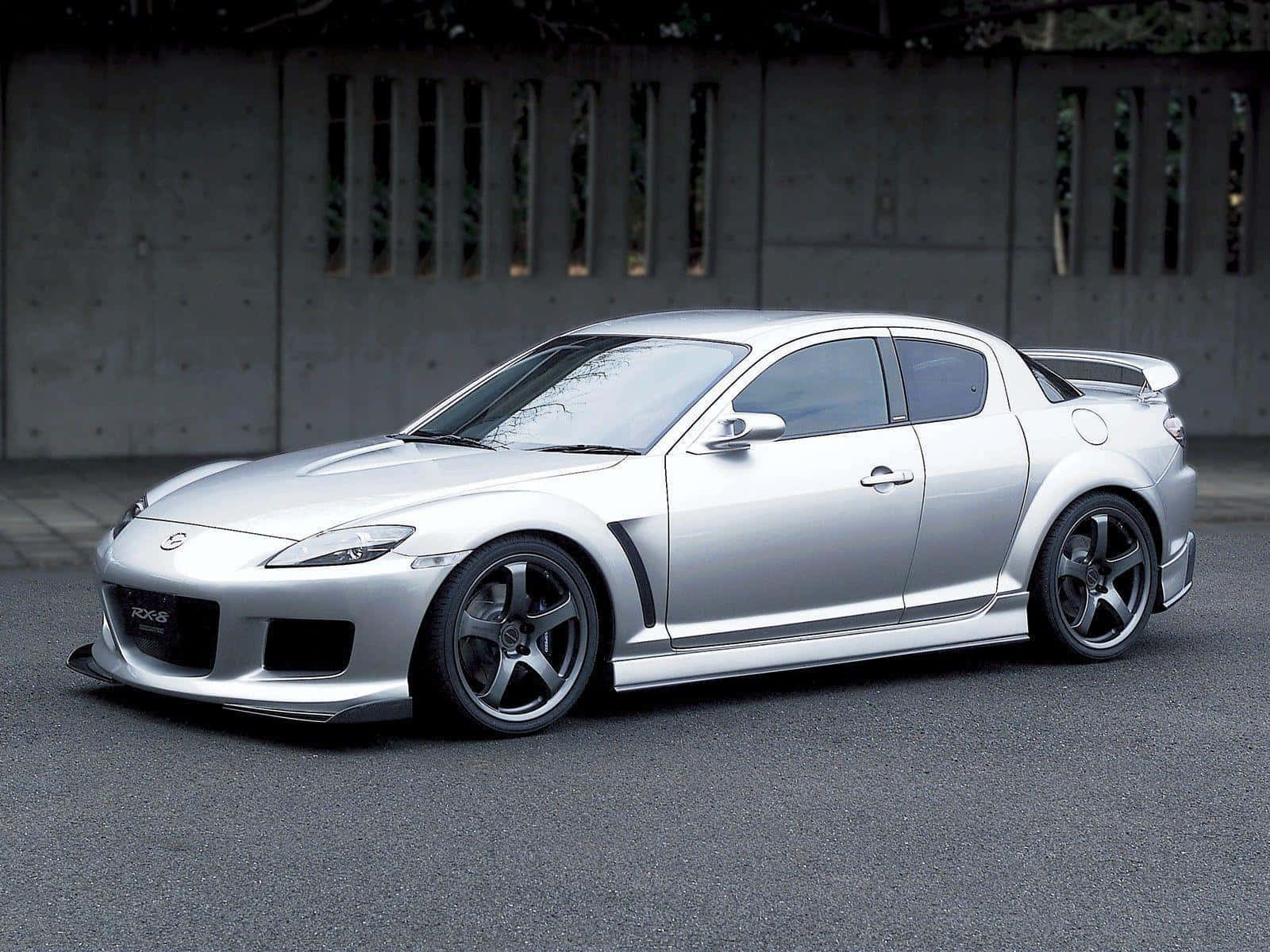 Mazda Rx-8 桌布 壁紙 — 一輛時尚的銀色馬自達RX-8跑車，配備黑色輪圈，停在柏油路面上，背景是帶有垂直縫隙的混凝土牆。