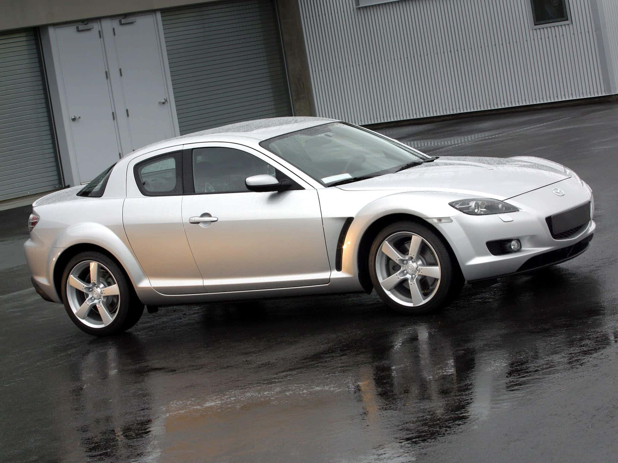 Mazda Rx-8 桌布 壁紙 — 一輛時尚的銀色馬自達RX-8轎跑車在濕瀝青路面上閃閃發光，其曲線倒映在灰色工業建築背景下。
