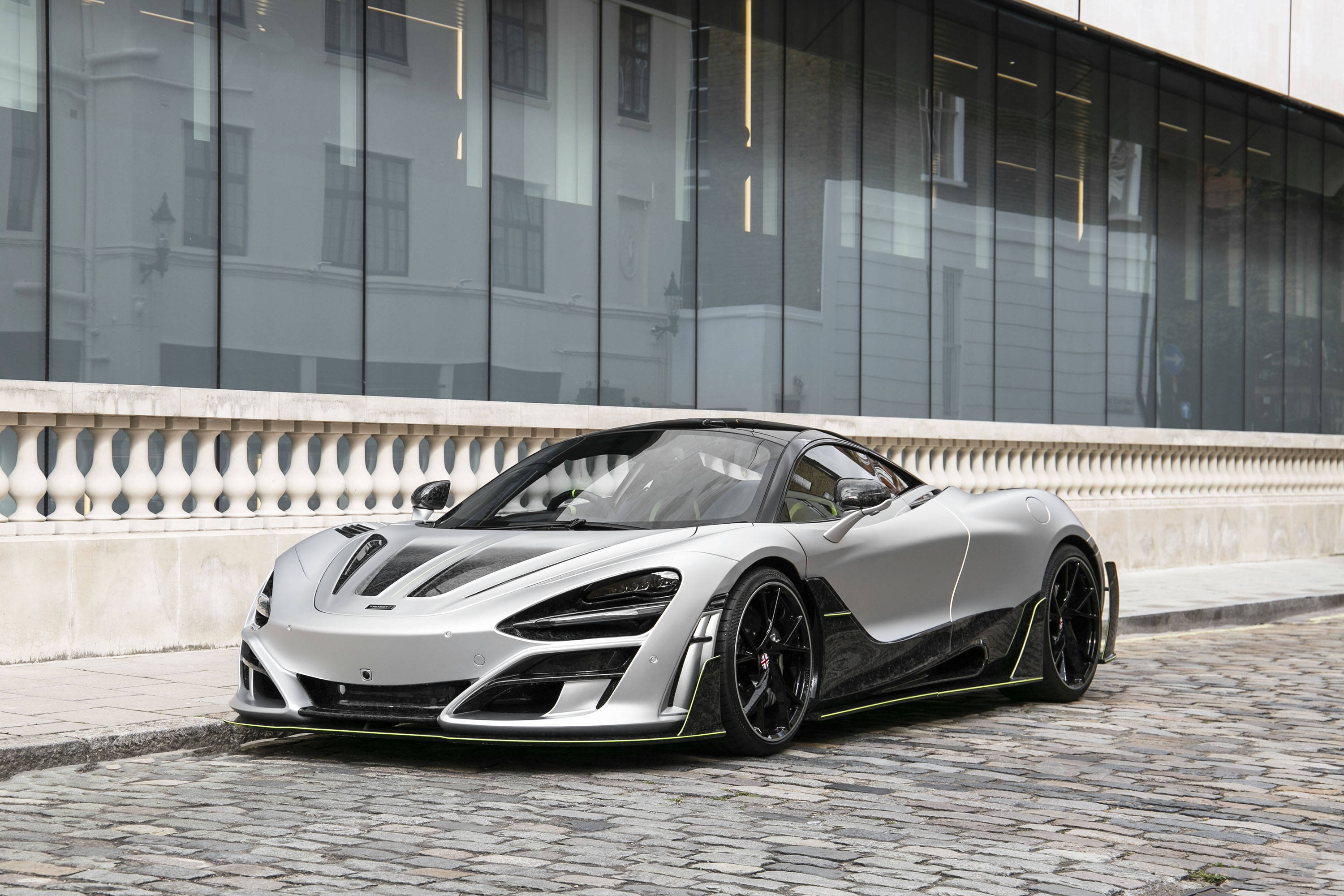 Mclaren 720s 桌布 壁紙 — 一輛時尚的銀色麥拉倫跑車，帶有黑色飾邊，停泊在現代玻璃建築旁的鵝卵石街道上。