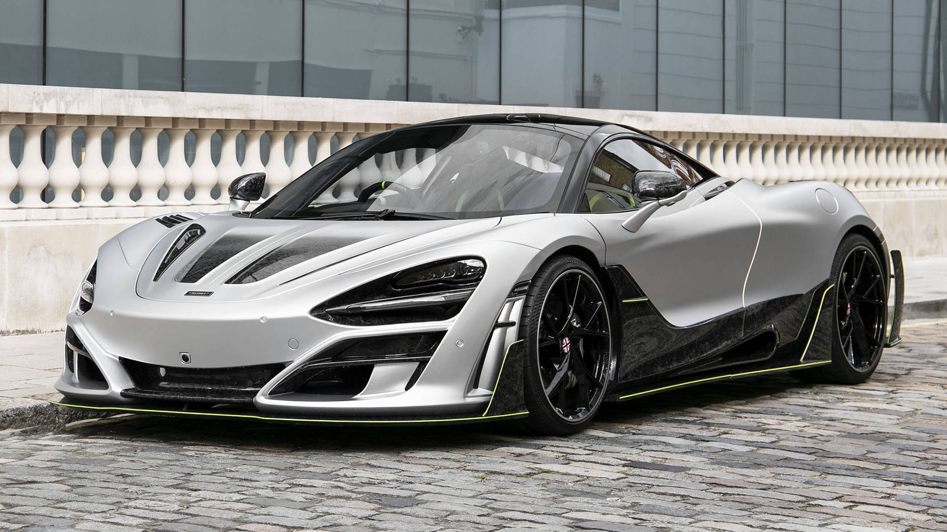 mclaren 720s 4K 桌布 壁紙 — 一輛時尚的銀色麥拿倫跑車，帶有黑色飾邊和螢光綠色裝飾，停在現代建築附近的鵝卵石街道上。