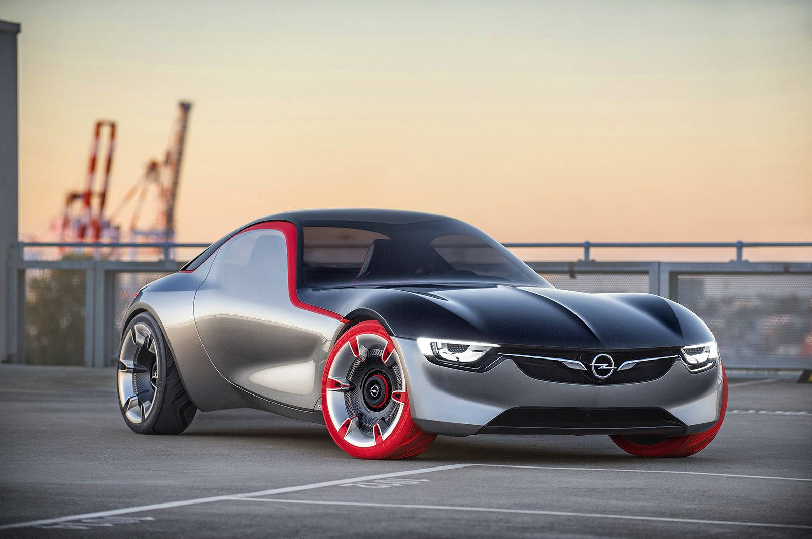 Opel 桌布 壁紙 — 一輛時尚的銀黑色概念車，帶有紅色點綴，停泊在碼頭上，背景是夕陽和起重機。