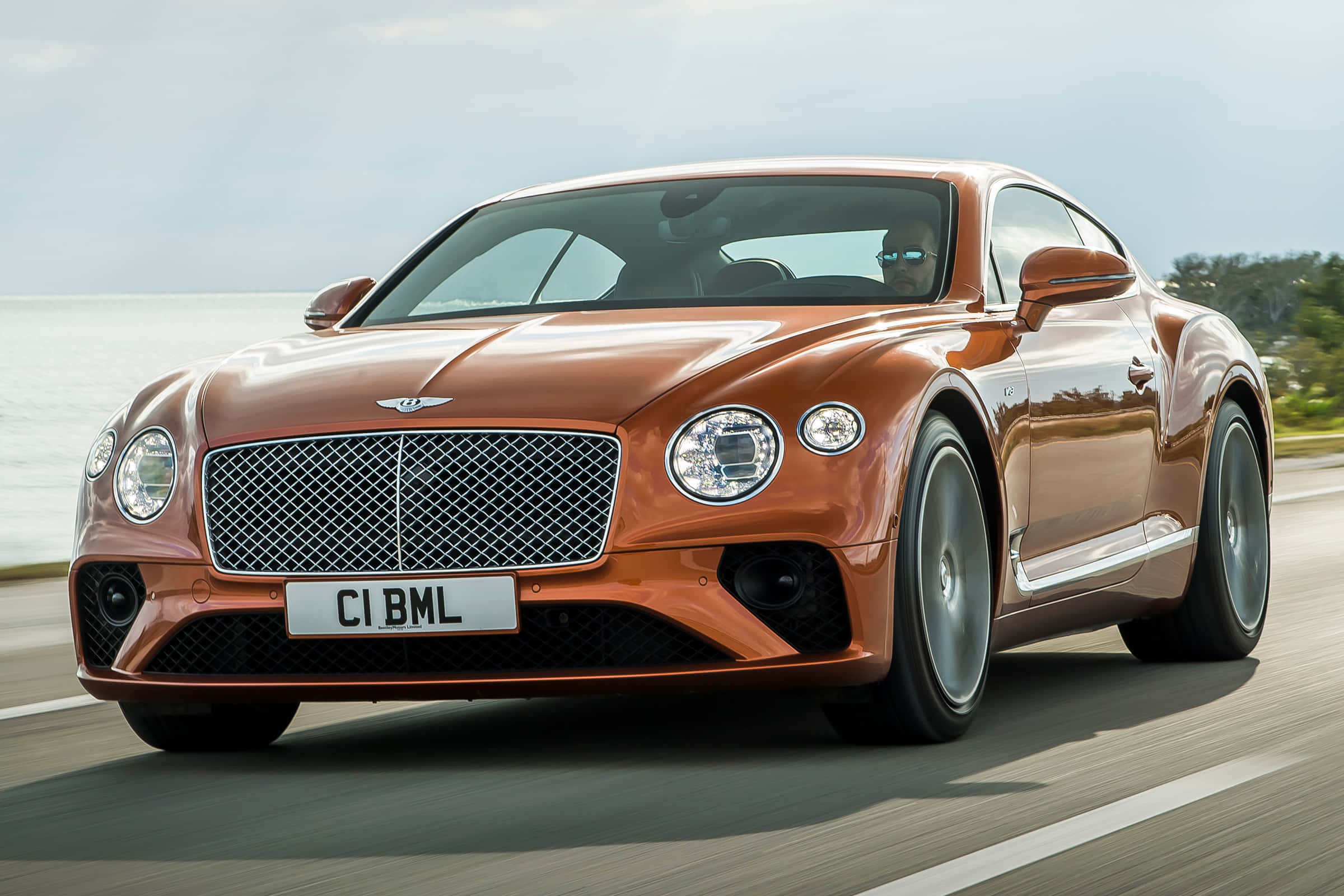 賓利 桌布 壁紙 — 一輛時尚的銅色 Bentley Continental GT 沿著海岸公路疾馳，背景是海洋和天空。