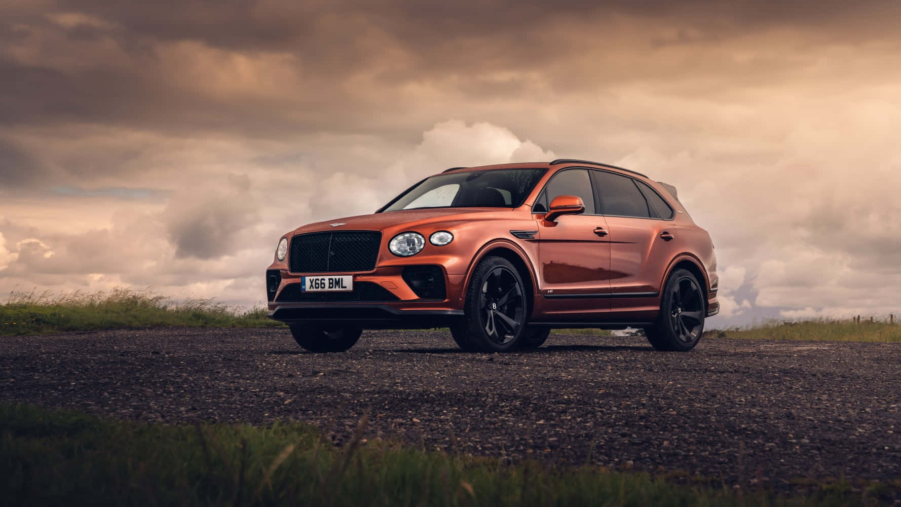 賓利 桌布 壁紙 — 一輛時尚的銅色賓利 Bentayga SUV 停泊在碎石路上，天空烏雲密佈，氣氛戲劇化。