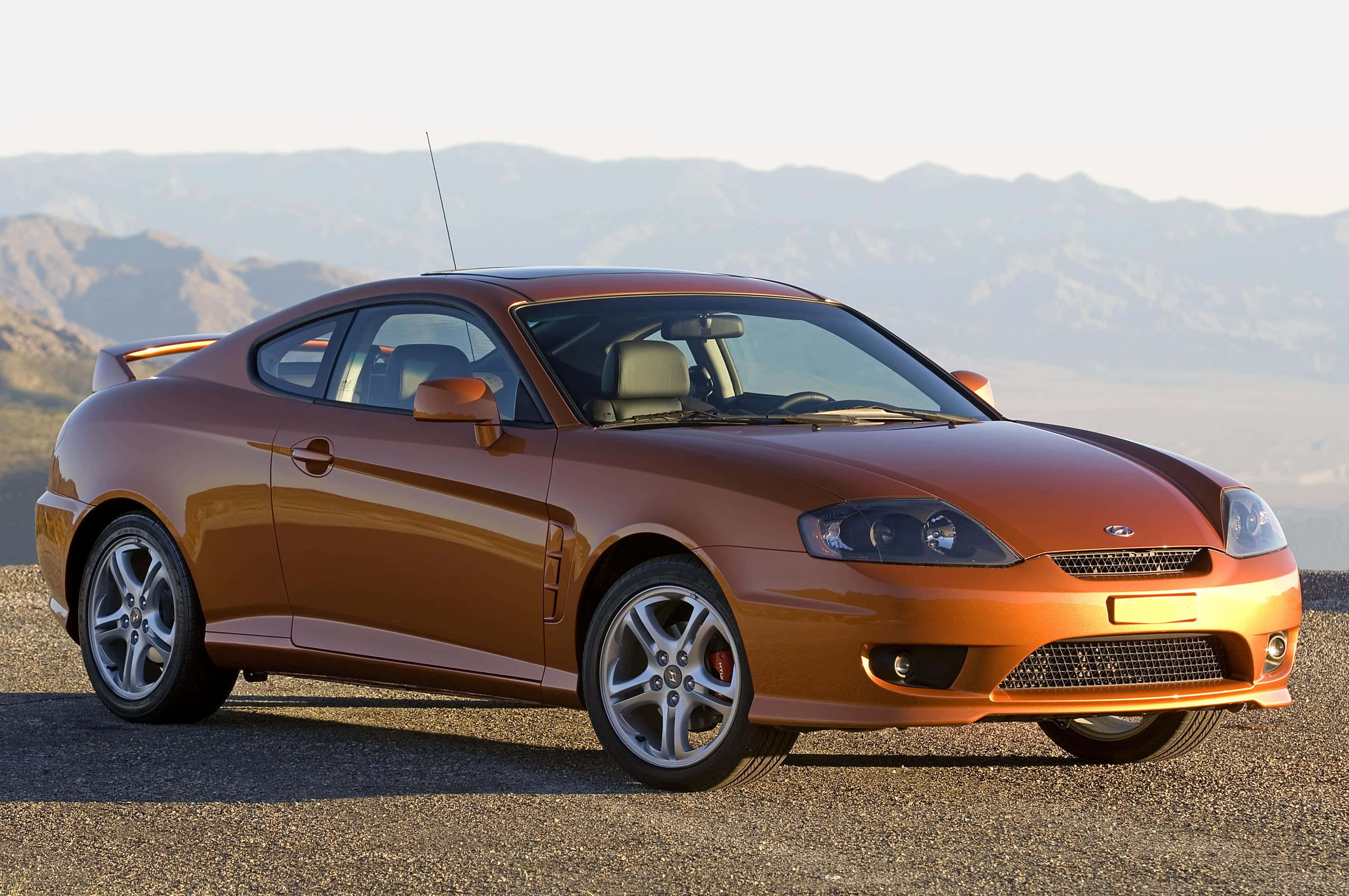 Hyundai Tiburon 桌布 壁紙 — 一輛時尚的銅色跑車停在碎石路上，背景是朦朧的山脈，在柔和的白日光線下。