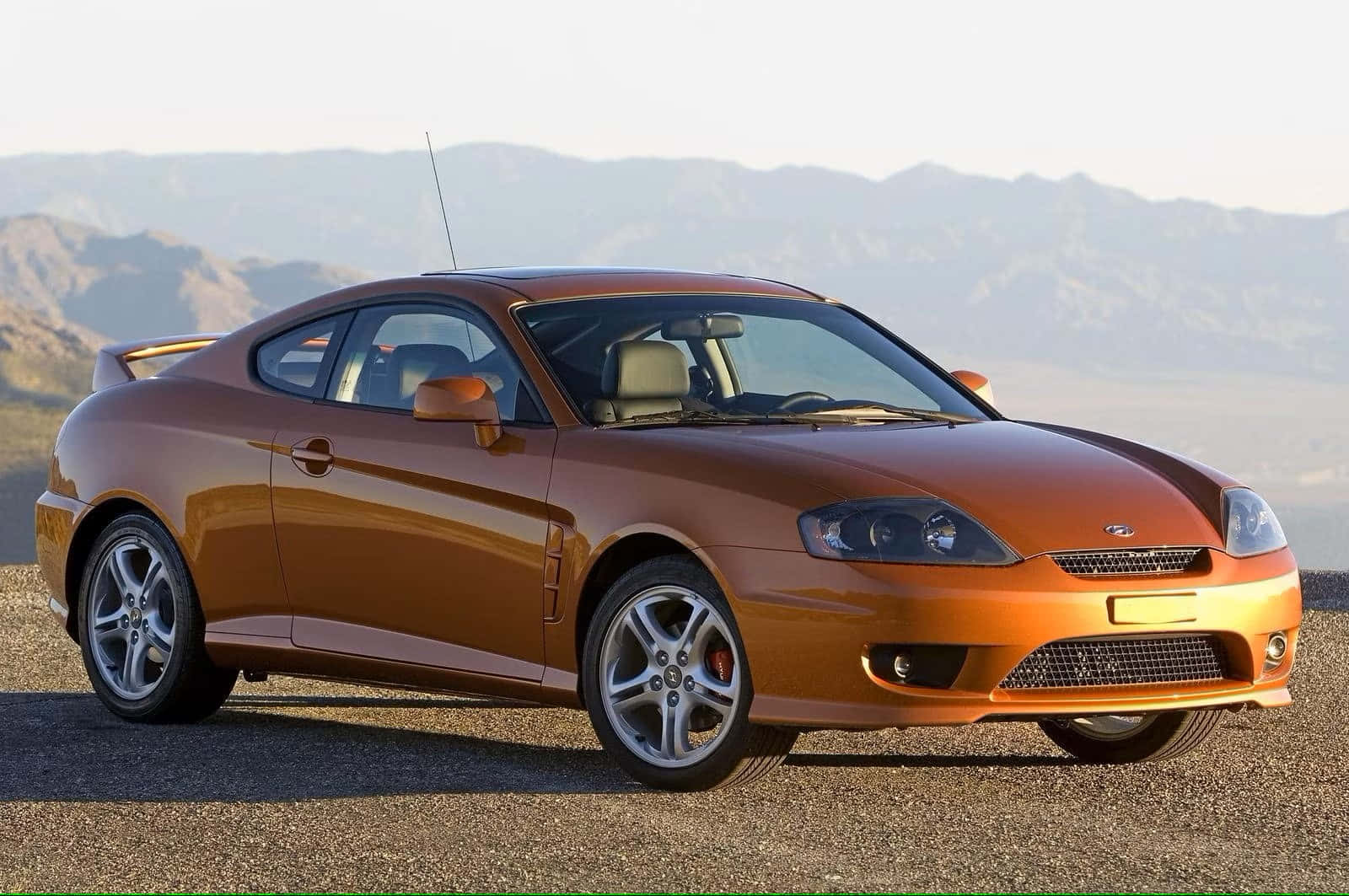 Hyundai Tiburon 桌布 壁紙 — 一輛時尚的銅色運動轎跑車停在碎石路上，背景是明亮的日光下朦朧的山脈。