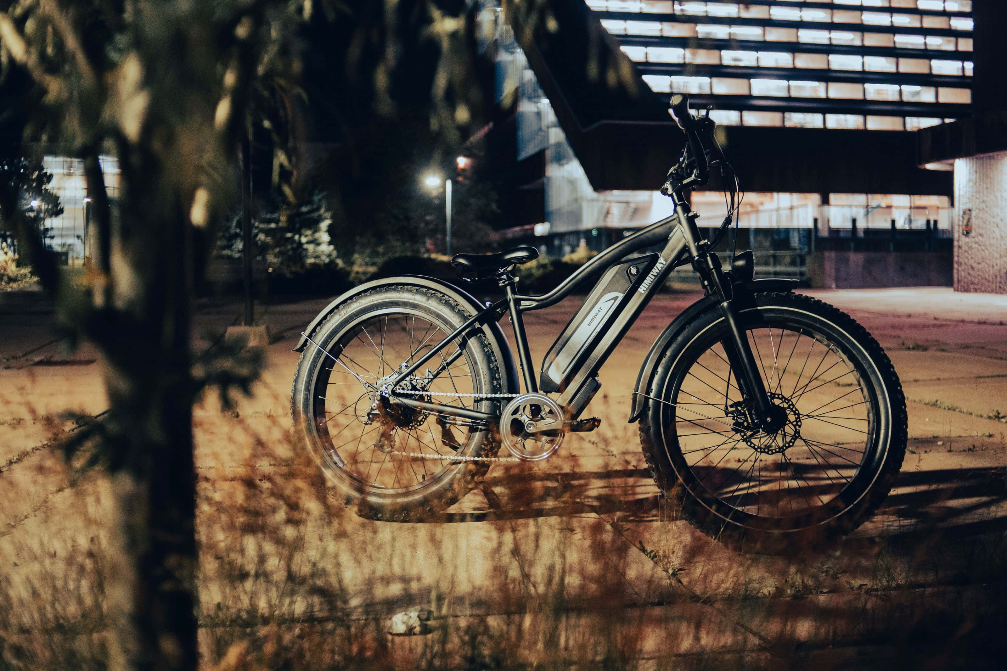 Electric Bike 桌布 壁紙 — 一輛時尚的電動自行車在夜間停放在現代建築下，溫暖的環境燈光投下長長的陰影。