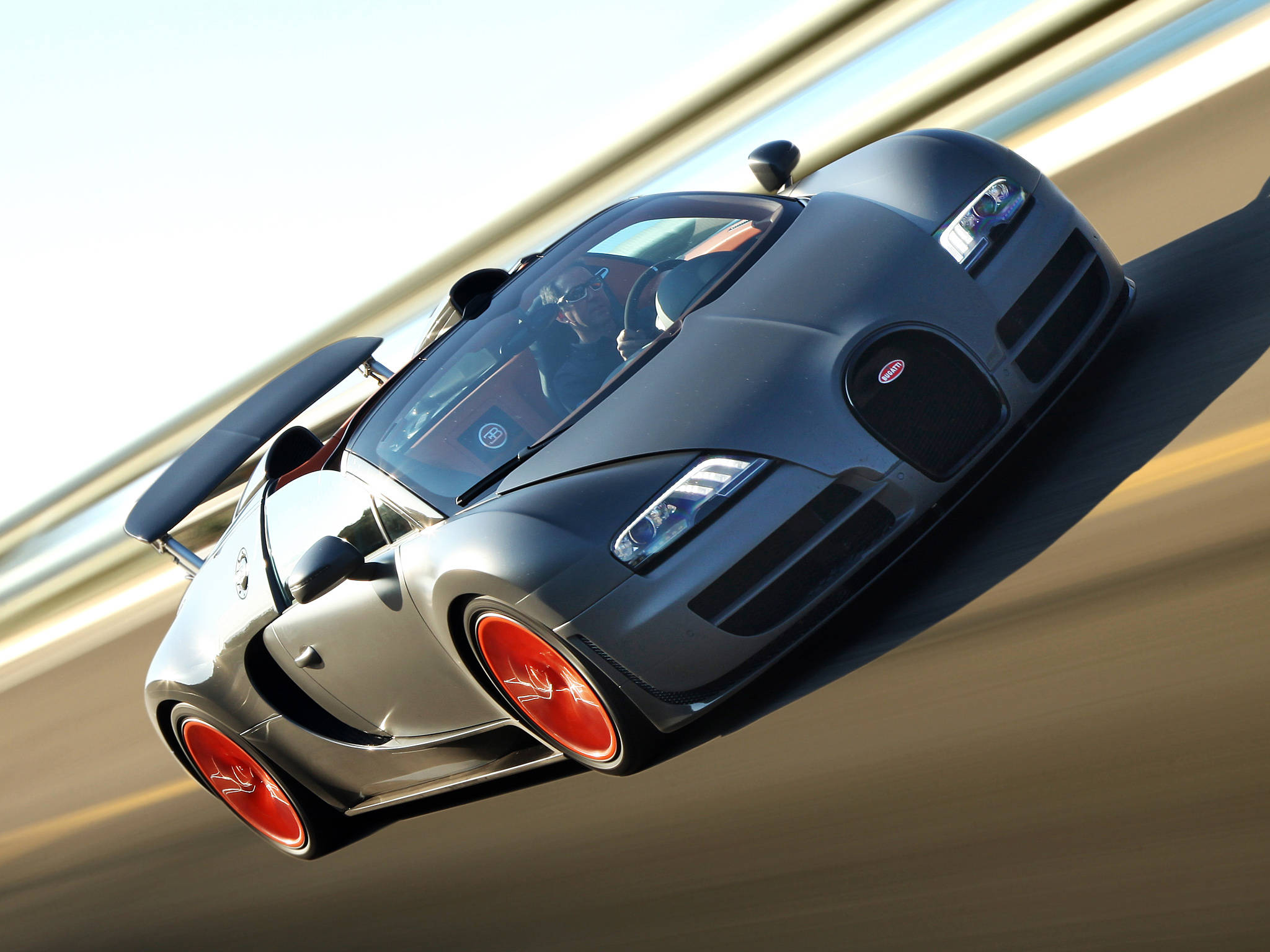 bugatti iPhone 桌布 壁紙 — 一輛時尚的霧面黑色Bugatti Veyron在模糊的賽道上疾馳，配備鮮紅色車輪，車內駕駛員清晰可見。