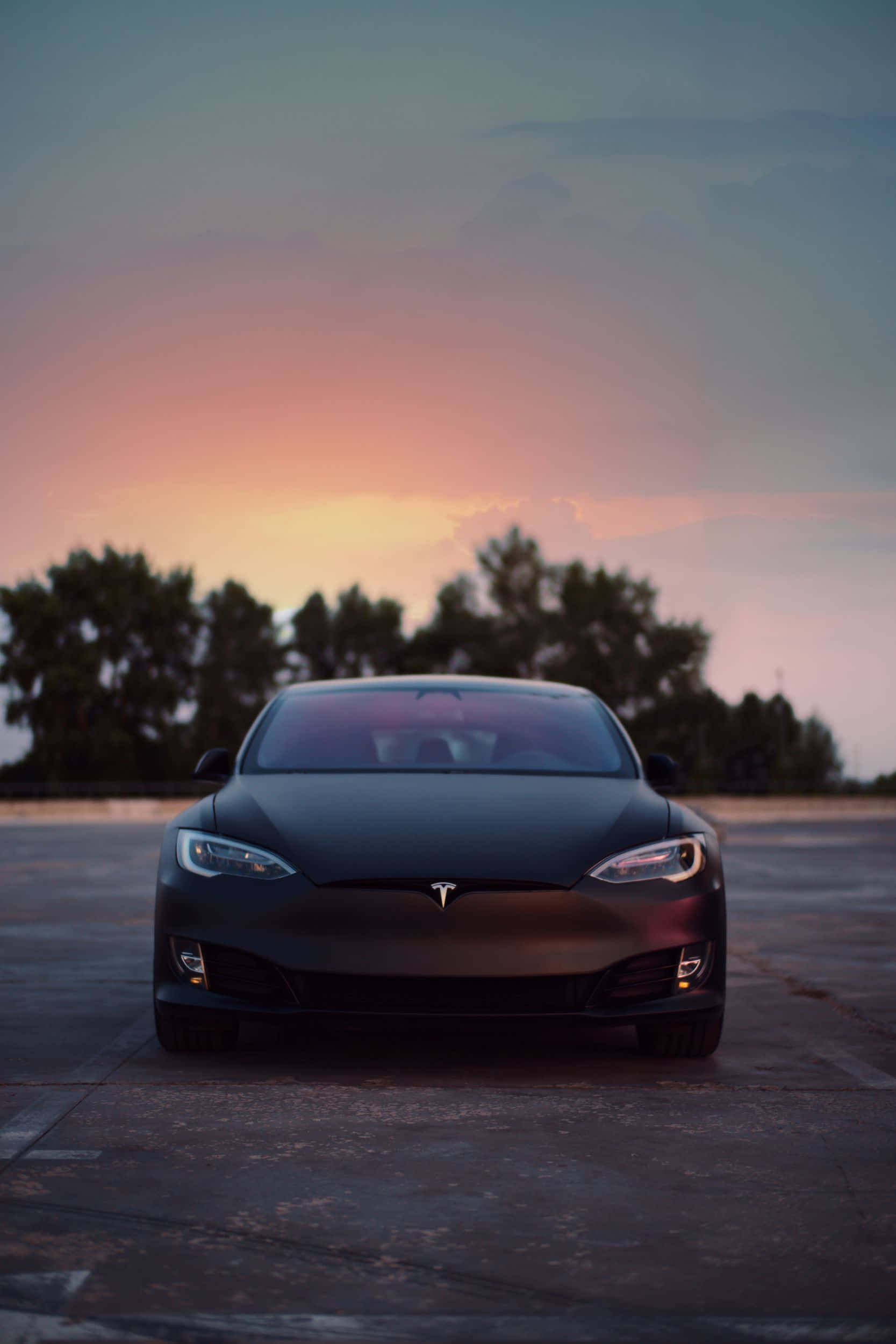 Hybrid Car 桌布 壁紙 — 一輛時尚的霧面黑色Tesla汽車在鋪砌的停車場上面向前方，背景是柔和的粉紅色和橙色日落天空，樹木形成剪影。