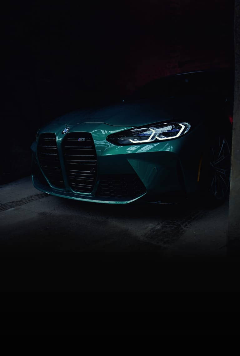 m 桌布 壁紙 — 一輛時尚的青綠色BMW M3在昏暗的車庫中被戲劇性的燈光照亮，突顯其激進的前格柵和發光的車頭燈。