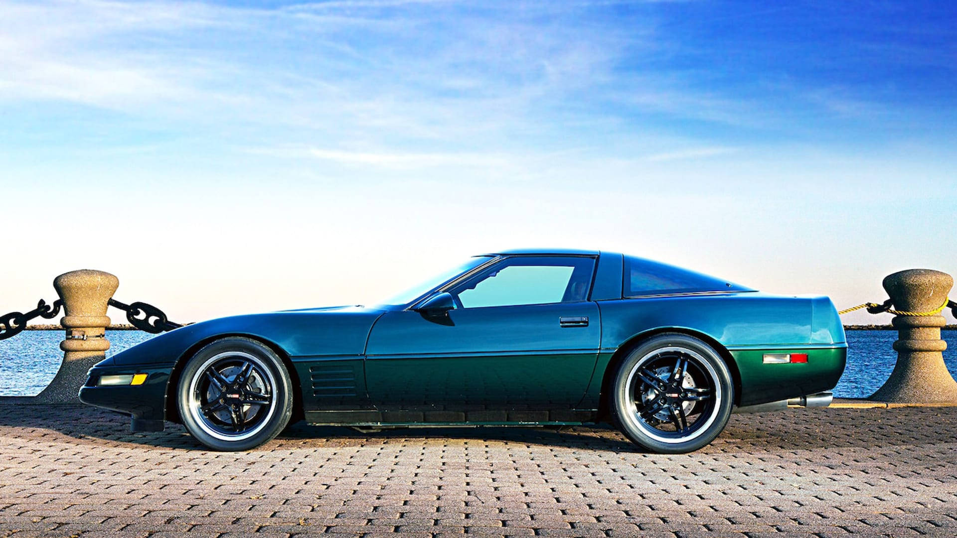 corvette 桌布 壁紙 — 一輛時尚的青綠色Corvette跑車停泊在平靜水面旁的鵝卵石碼頭，天空晴朗蔚藍。