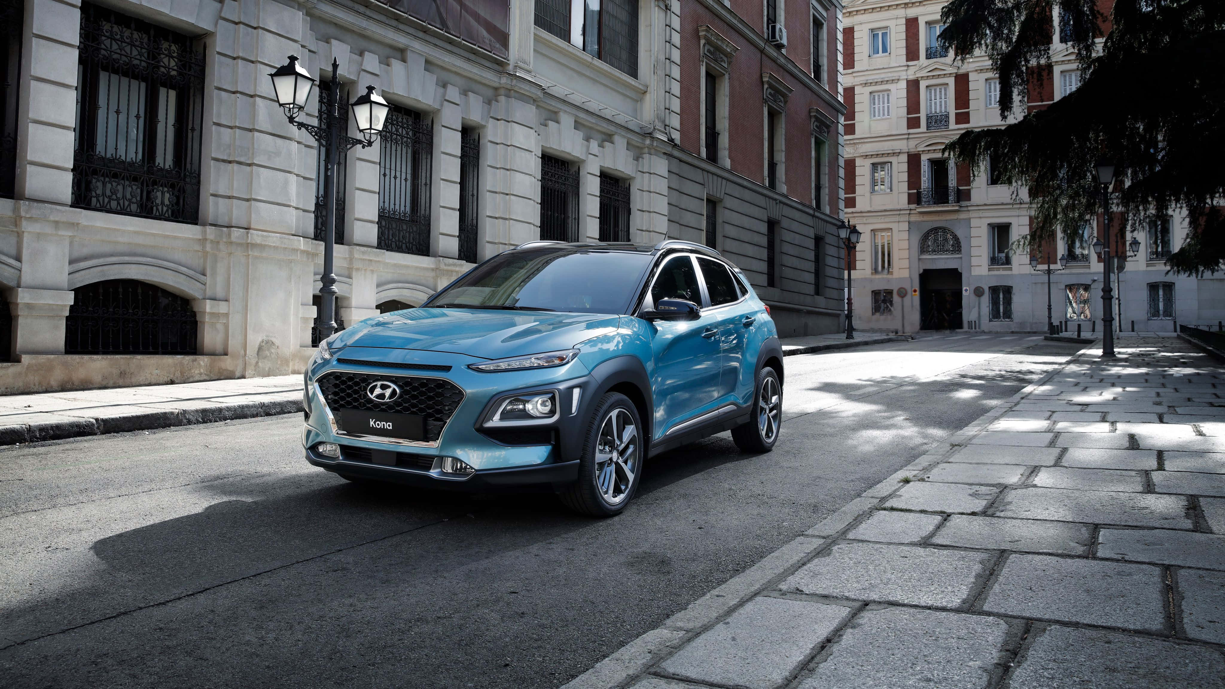 Hyundai Kona 桌布 壁紙 — 一輛時尚的青綠色Hyundai Kona SUV停放在陽光普照的鵝卵石街道上，旁邊是歷史悠久的歐洲建築。