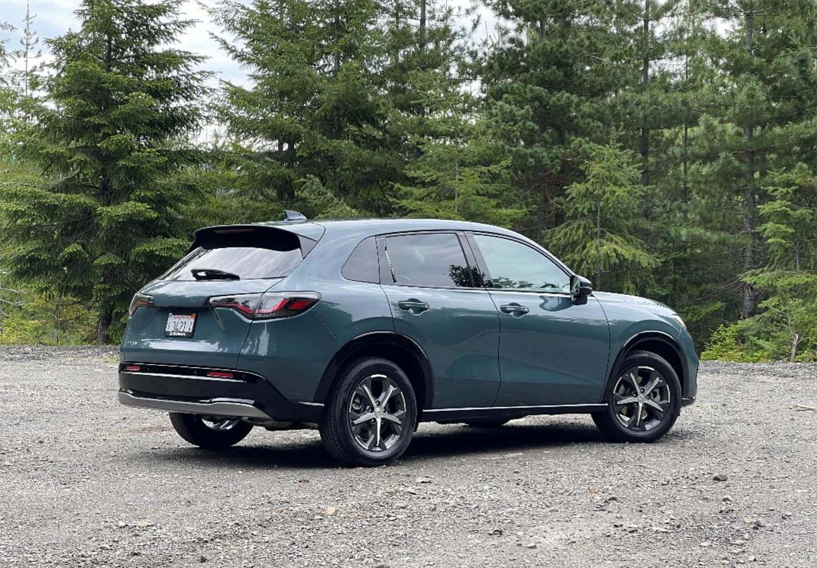 Honda Hr-v 桌布 壁紙 — 一輛時尚的青綠色SUV停泊在碎石上，背景是高聳的常青樹，天空多雲。
