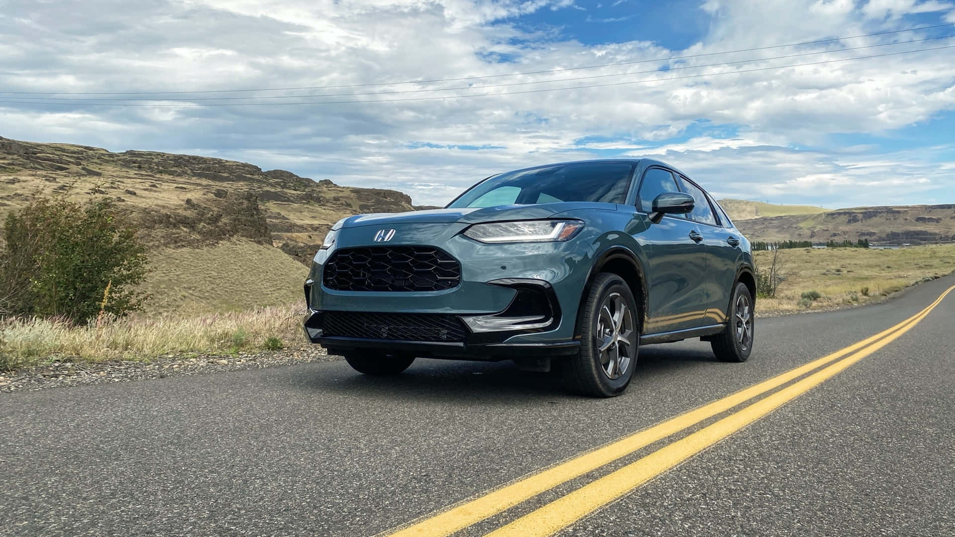 Honda Hr-v 桌布 壁紙 — 一輛時尚的青綠色SUV沿著蜿蜒的沙漠道路行駛，天空部分多雲，背景是起伏的丘陵。