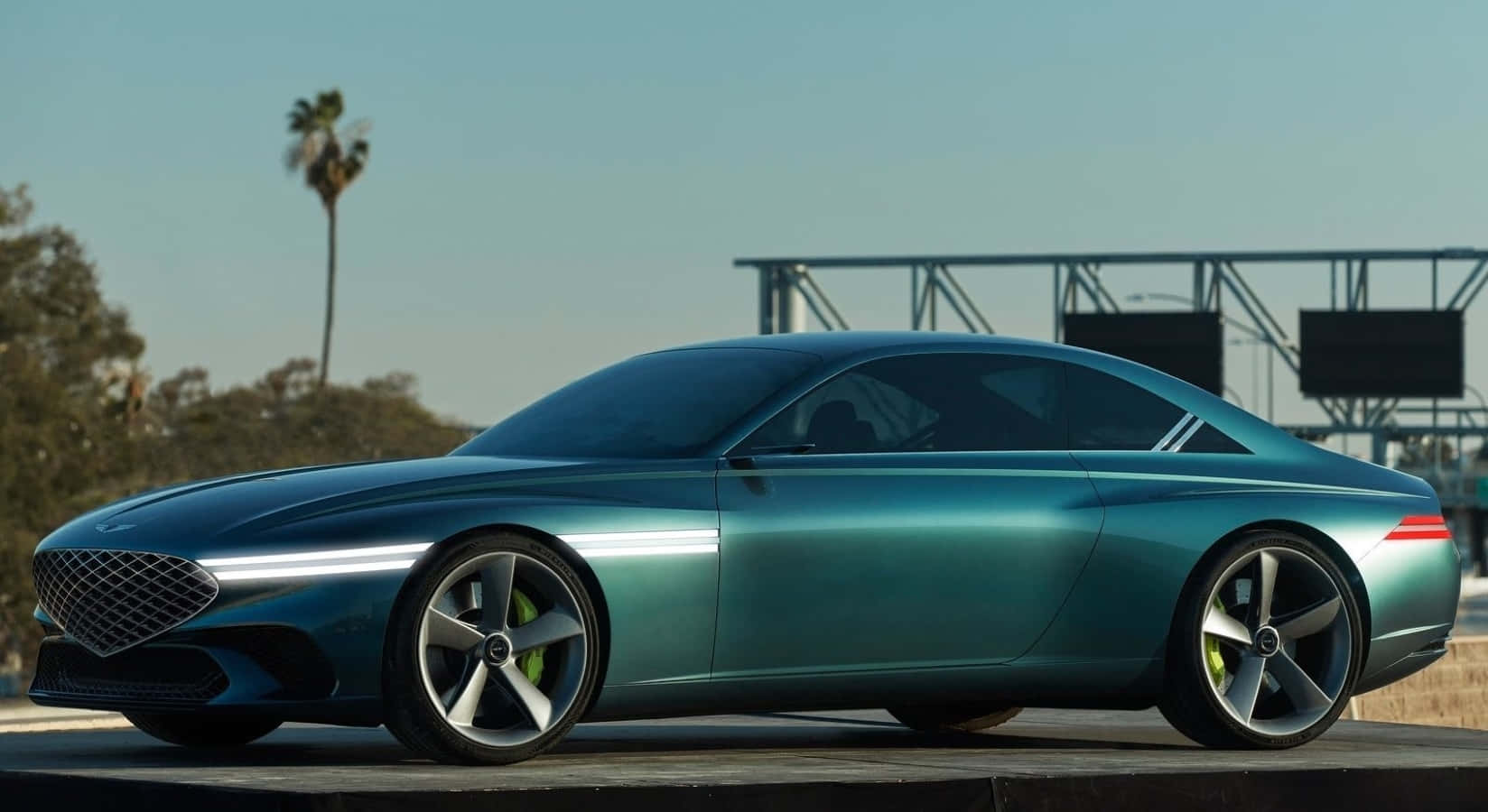 Genesis X Concept 桌布 壁紙 — 一輛時尚的青綠色概念車，帶有發光裝飾，停放在平台上，背景是晴朗的天空、棕櫚樹和體育場建築。