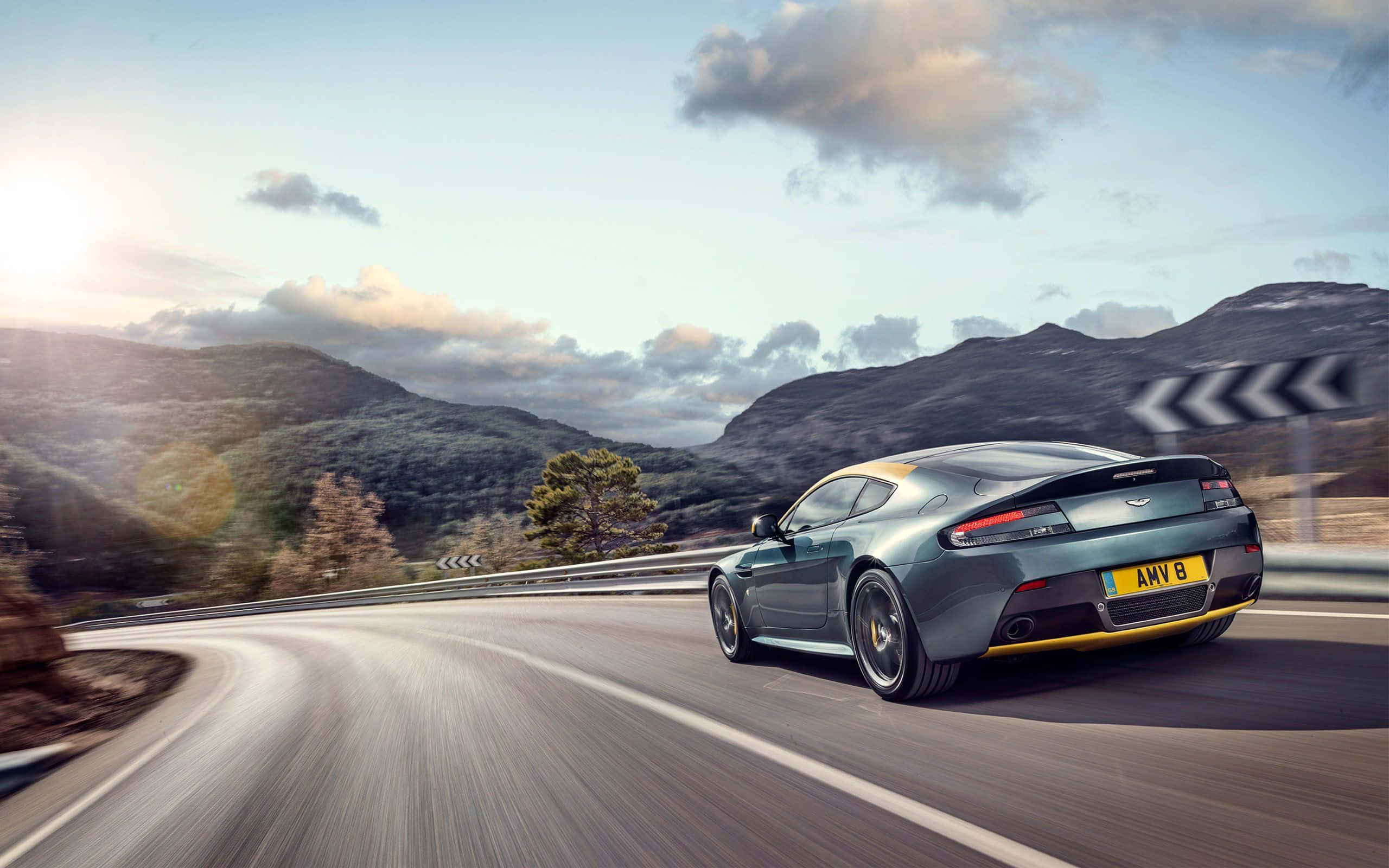 Aston Martin V12 Vantage 桌布 壁紙 — 一輛時尚的青綠色阿斯頓馬丁在明亮的陽光和散雲下沿著蜿蜒的山路飛馳。