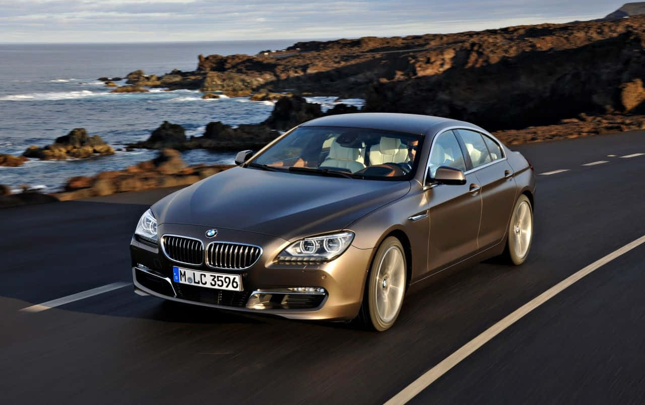 Bmw 6 Series 桌布 壁紙 — 一輛時尚的青銅色BMW 6系列轎跑車沿著海岸公路行駛，背景是崎嶇的懸崖和拍岸的浪花。