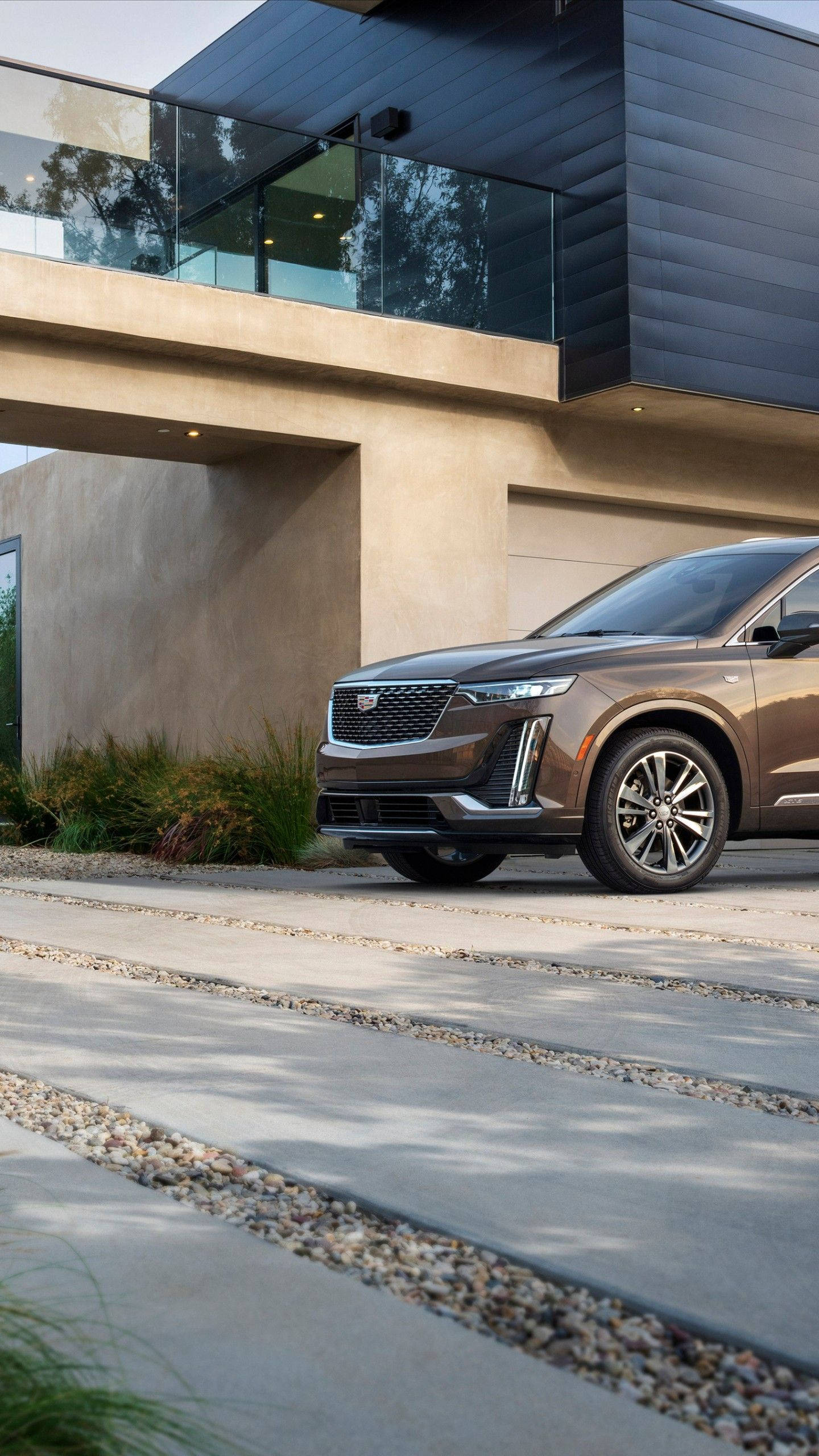 cadillac iPhone 桌布 壁紙 — 一輛時尚的青銅色SUV停放在現代混凝土車道上，前方是一座極簡風格的當代住宅，配有玻璃欄杆和紋理牆壁。