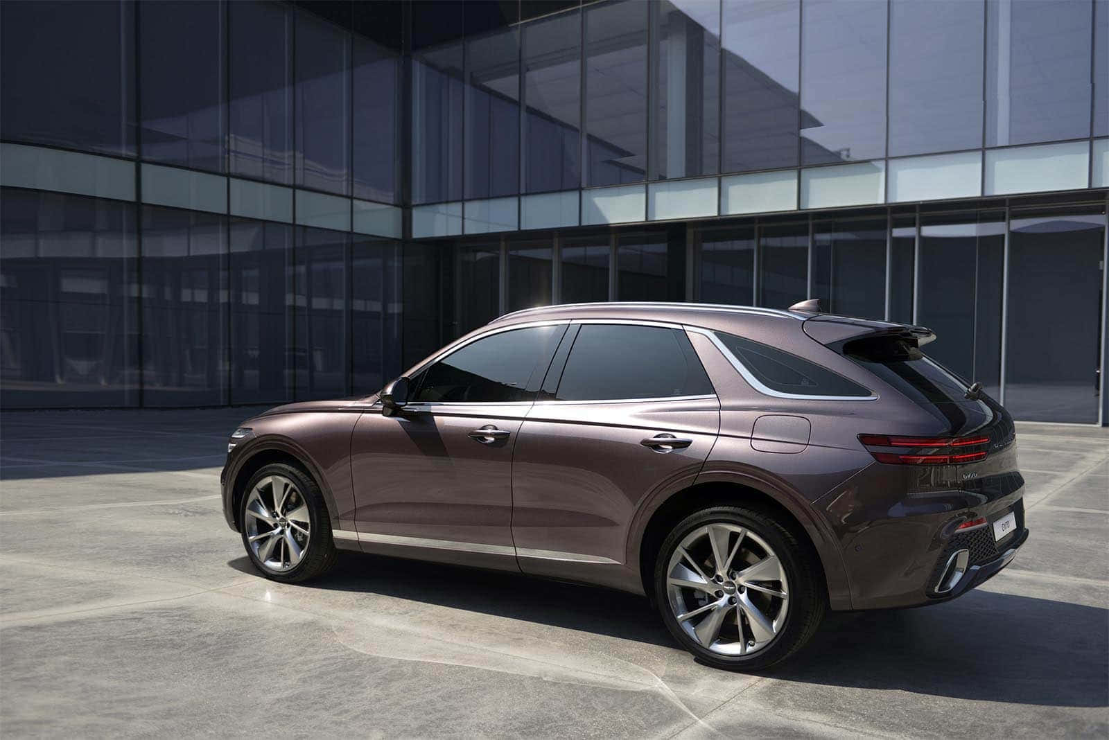 Genesis Gv70 桌布 壁紙 — 一輛時尚的青銅色SUV在明亮的日光下停放在現代玻璃建築前的混凝土上。