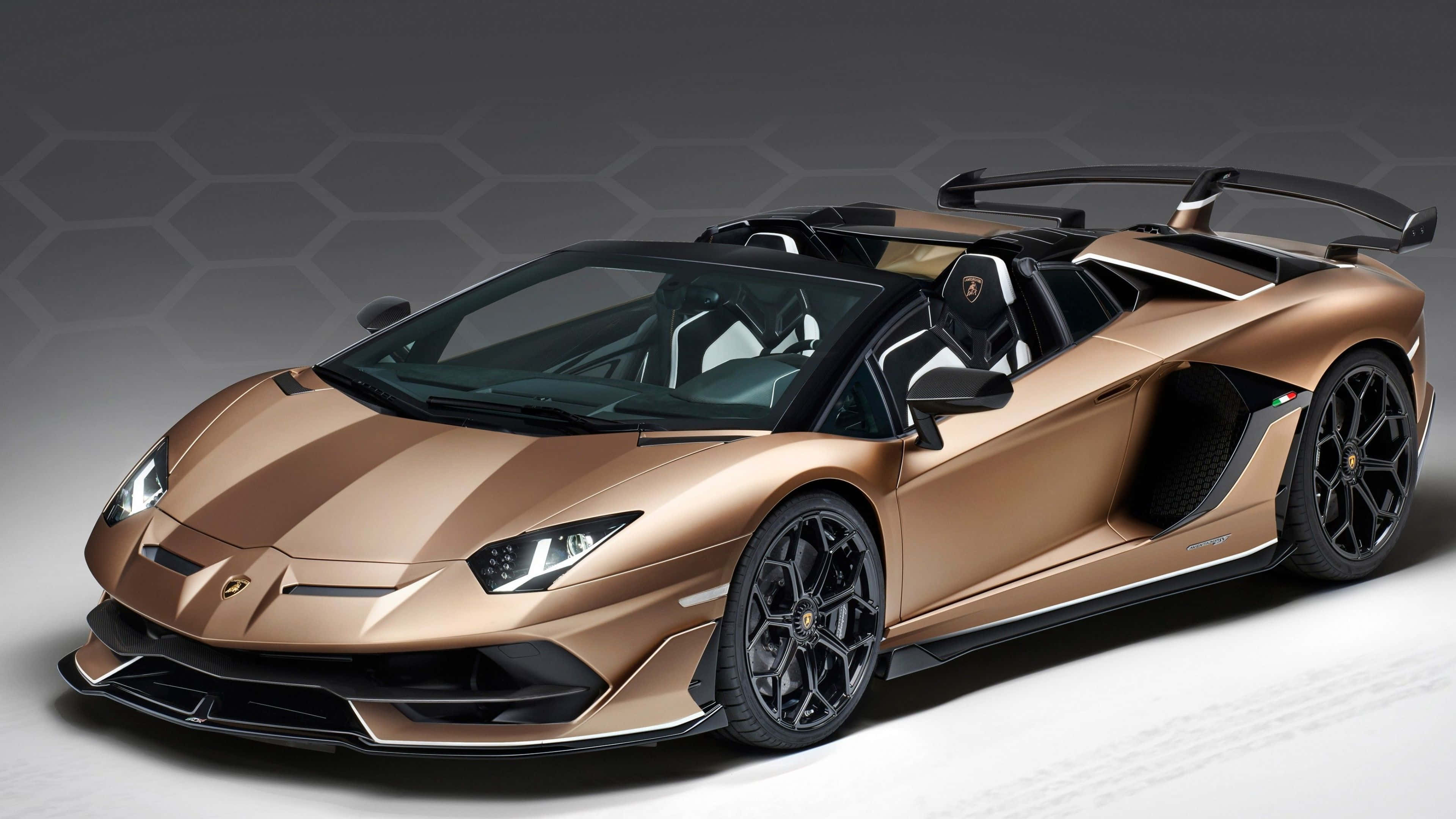 4K 藍寶堅尼 桌布 壁紙 — 一輛時尚的青銅色藍寶堅尼 Aventador SVJ Roadster，帶有黑色飾邊和突出的尾翼，在六邊形圖案的灰色工作室背景前擺拍。