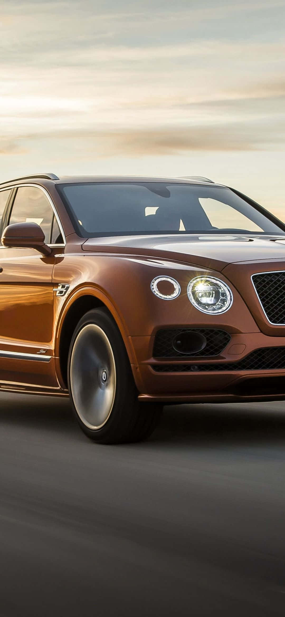 Bentley Cars 桌布 壁紙 — 一輛時尚的青銅色賓利Bentayga SUV在柔和的金色日落天空下，在模糊的道路上向前滑行。