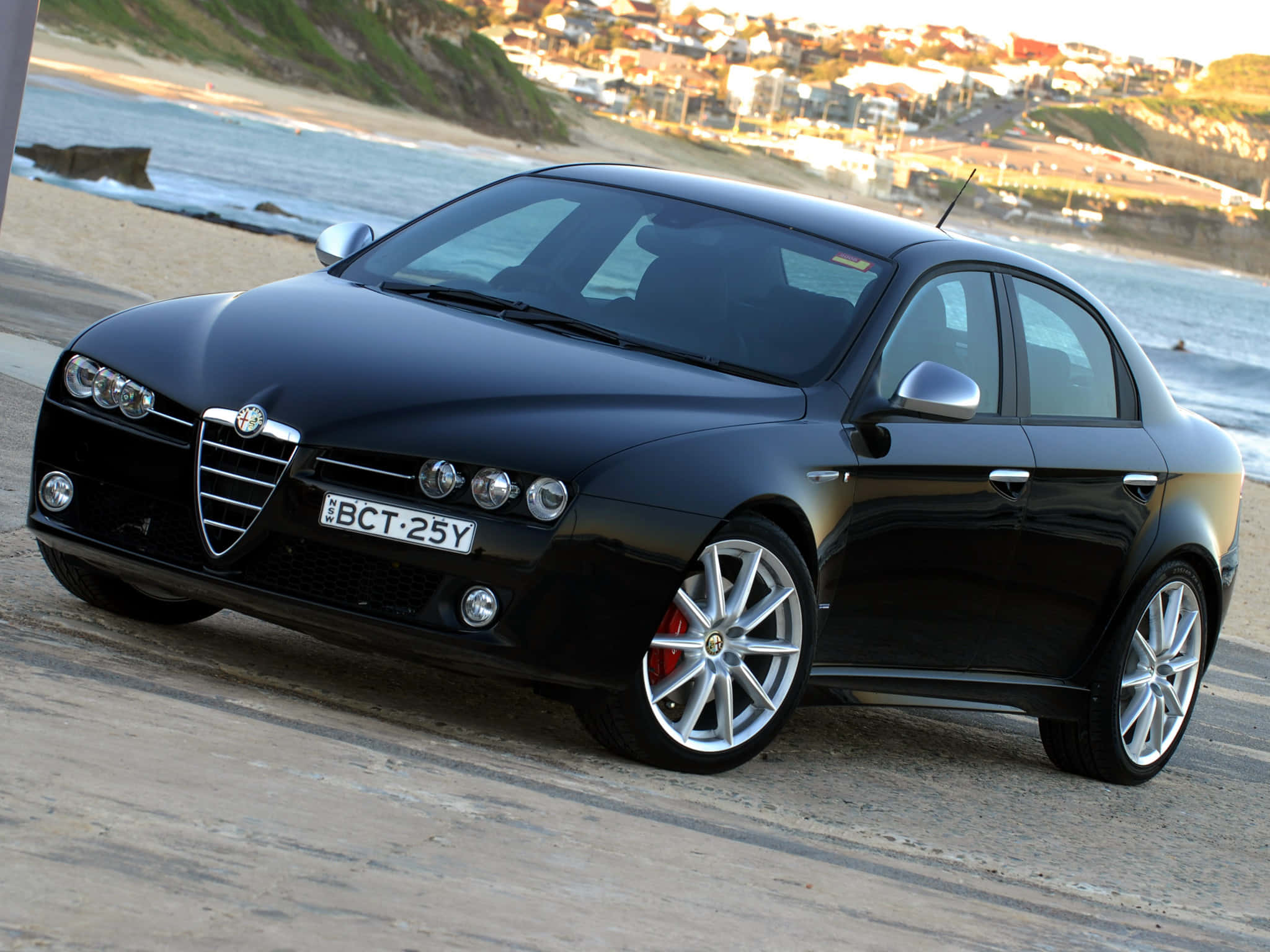 Alfa Romeo 159 桌布 壁紙 — 一輛時尚的黑色 Alfa Romeo 轎車停泊在沿海公路上，背景是海洋和懸崖小鎮。