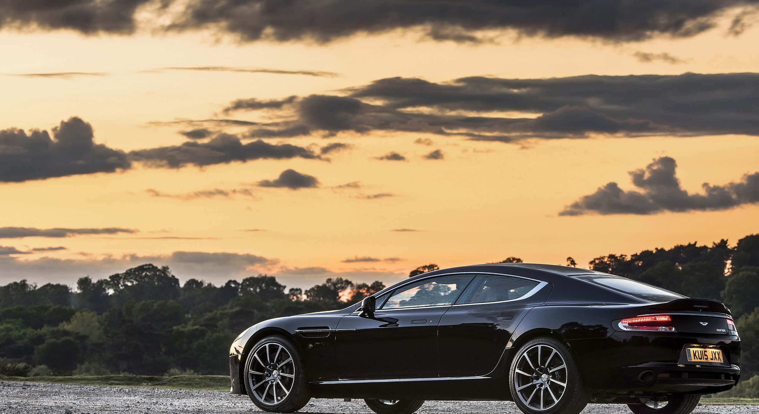 Aston Martin Rapide S 桌布 壁紙 — 一輛時尚的黑色 Aston Martin Rapide 停在夕陽下的碎石路上，上方有戲劇性的橙色和灰色雲彩。
