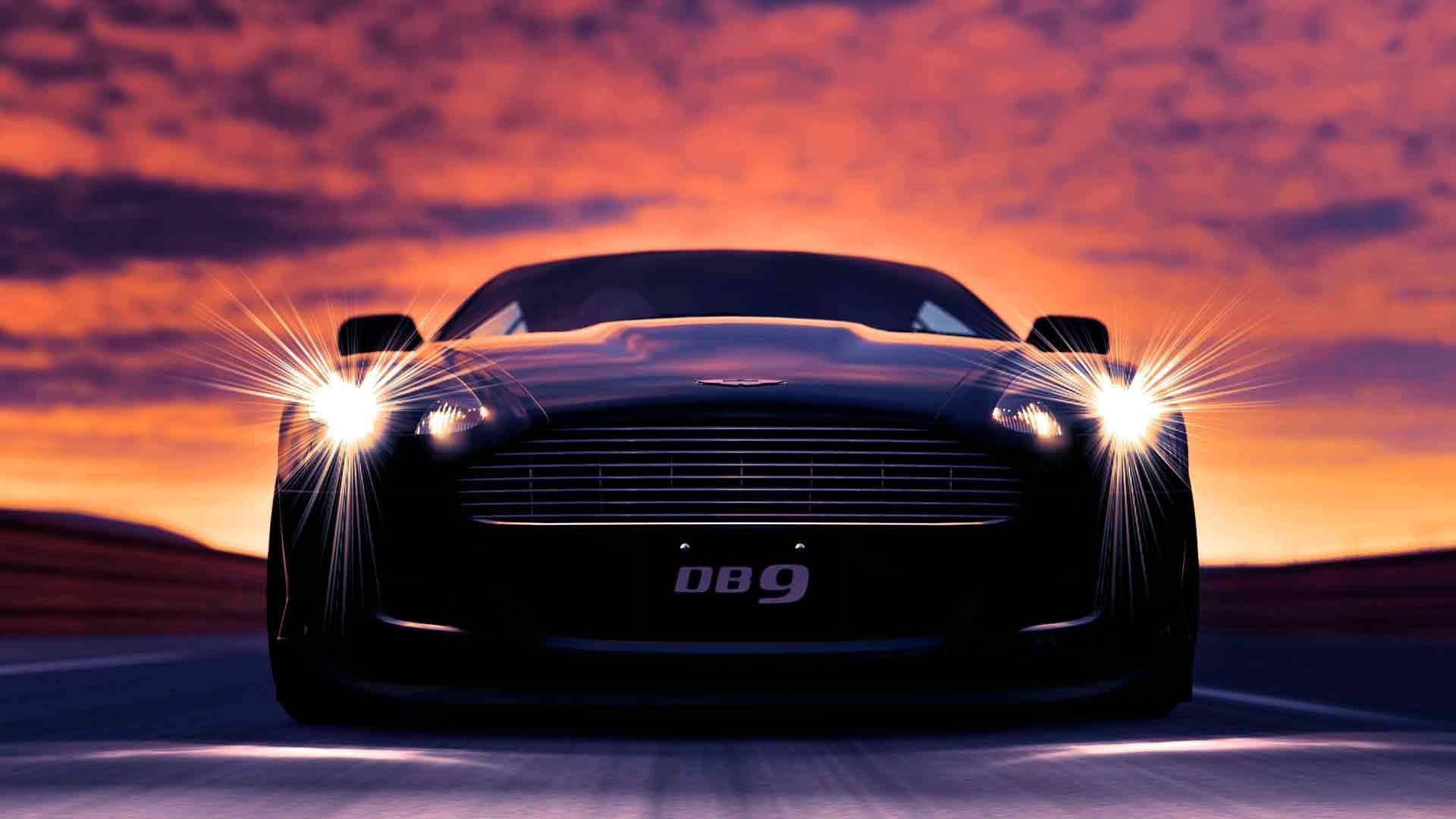 奧斯頓馬丁 桌布 壁紙 — 一輛時尚的黑色 Aston Martin DB9 在熾熱的夕陽天空下，亮著車頭燈向觀眾疾馳而來。
