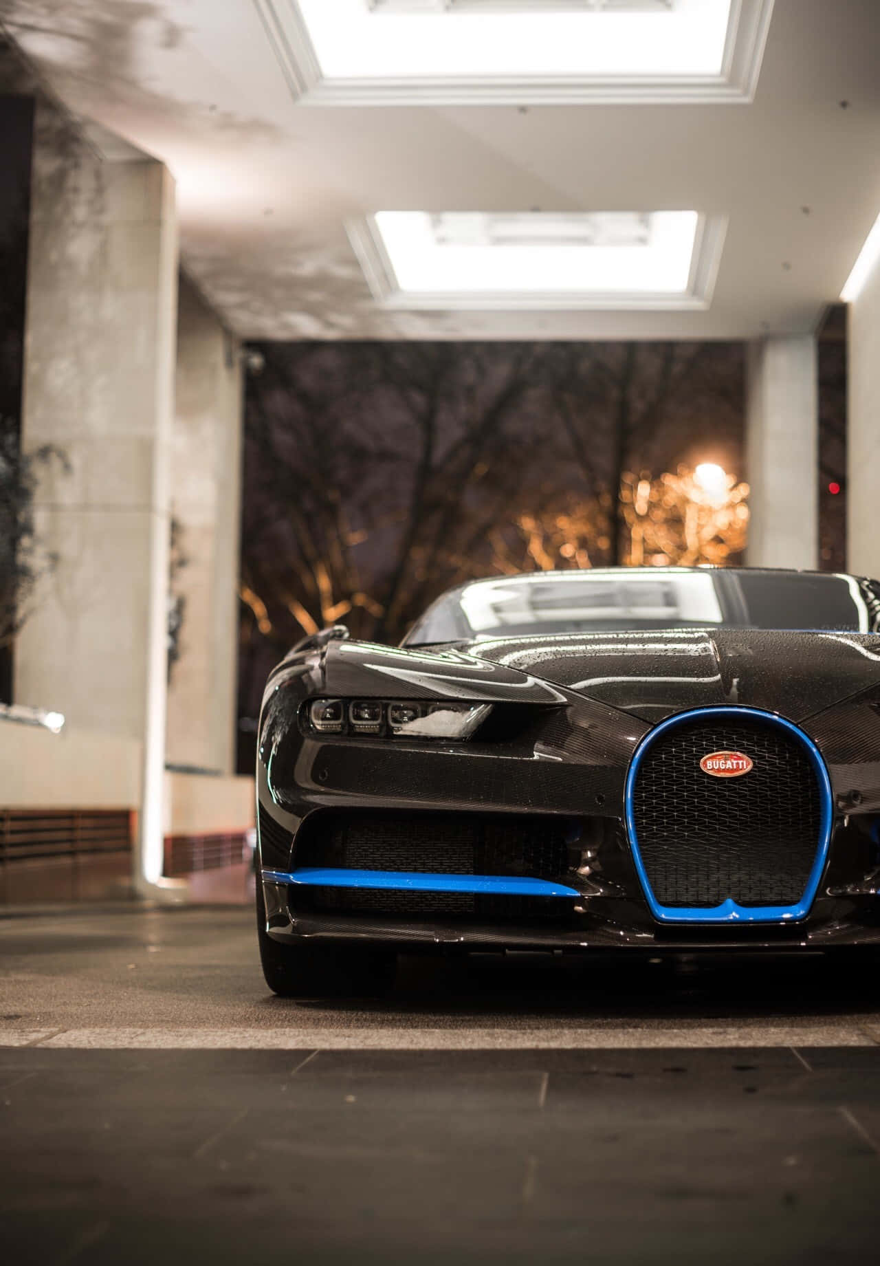 Bugatti Phone 桌布 壁紙 — 一輛時尚的黑色 Bugatti 超級跑車帶有發光的藍色飾邊，夜晚停放在現代建築的天窗下。