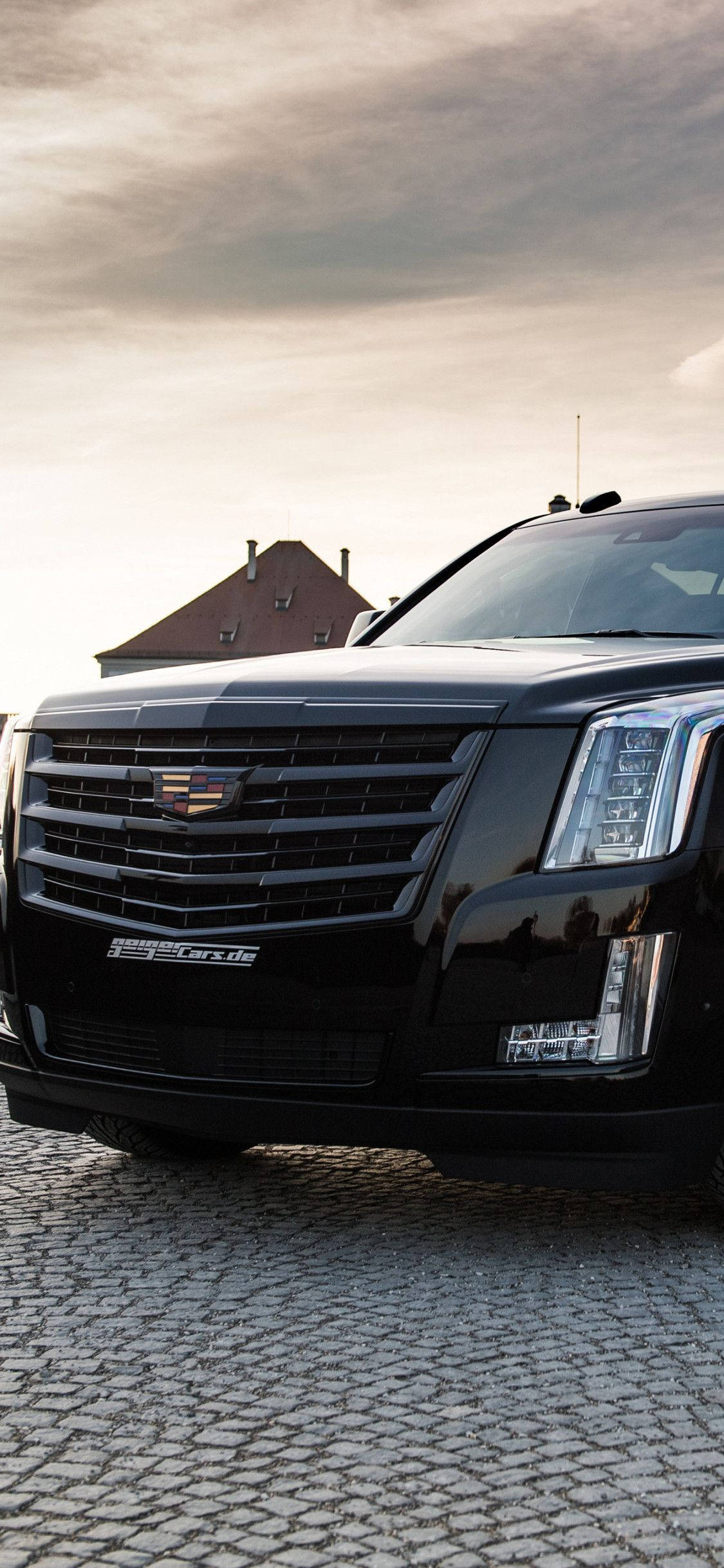 凱迪拉克 iPhone 桌布 壁紙 — 一輛時尚的黑色 Cadillac Escalade 停泊在鵝卵石街道上，天空多雲，背景有房屋。