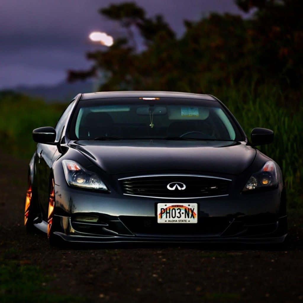 Infiniti G35 桌布 壁紙 — 一輛時尚的黑色 Infiniti 跑車，配備發光的橙色輪圈，停在土路上，天空暮色中有一輪明亮的月亮。