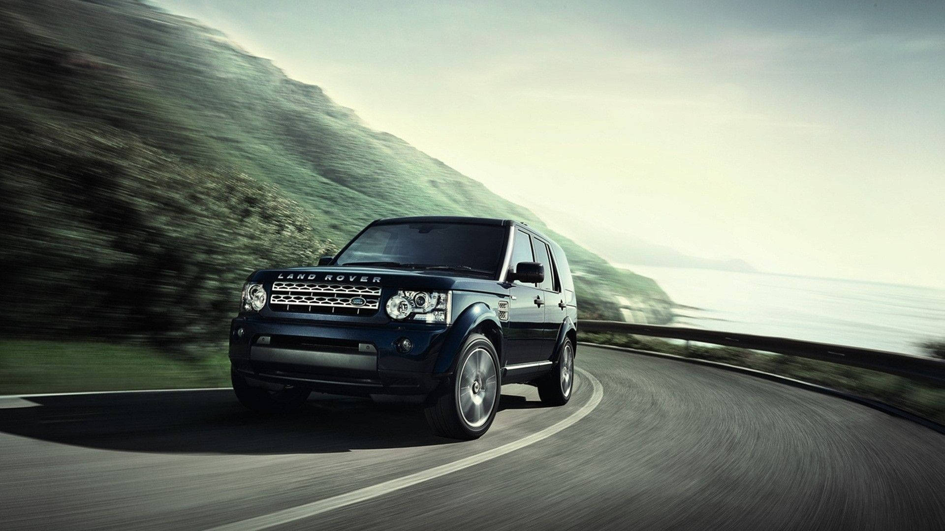 荒原路華 桌布 壁紙 — 一輛時尚的黑色 Land Rover 在蜿蜒的海岸道路上行駛，背景是模糊的綠色山丘和海洋。