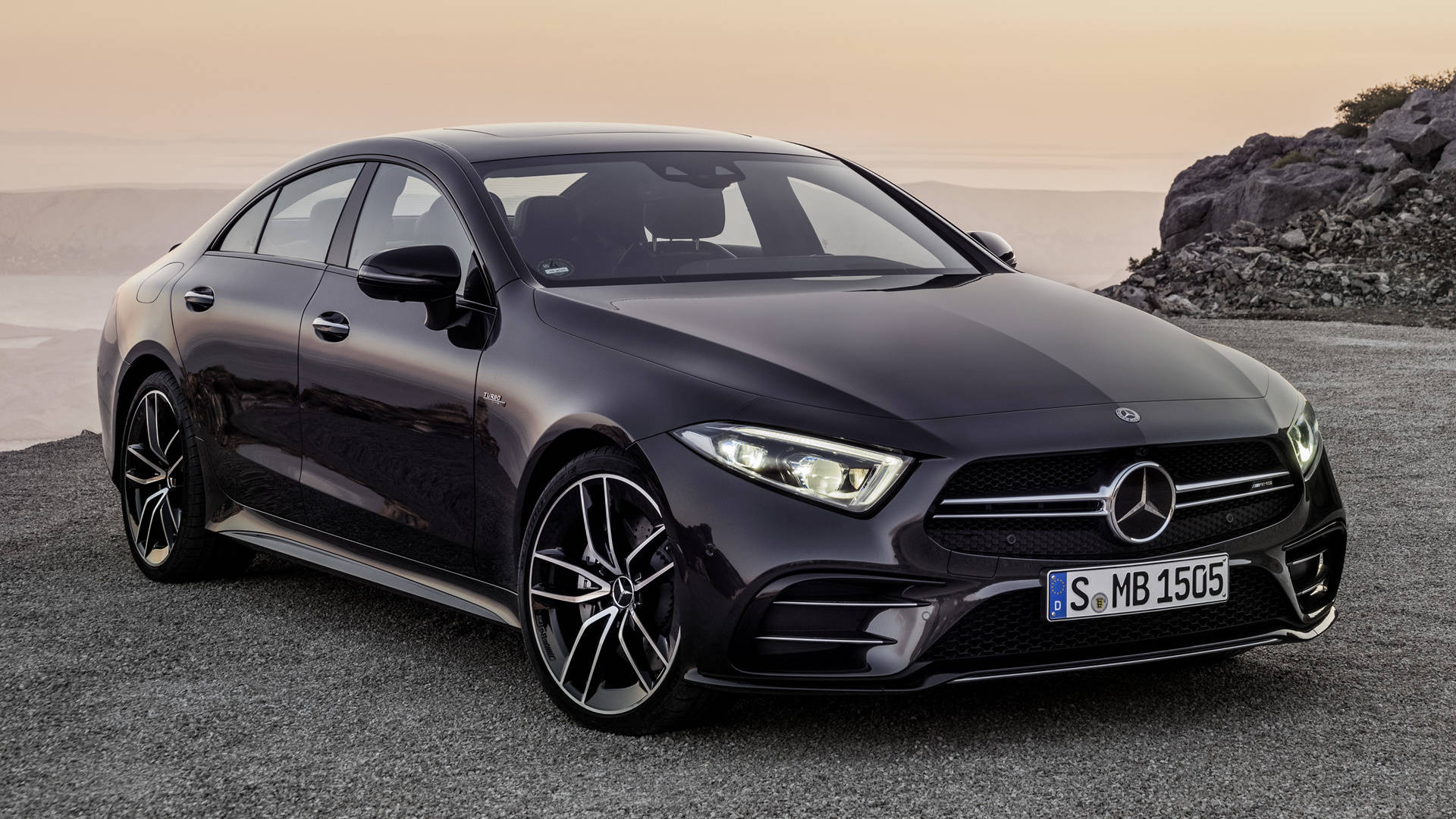Mercedes Amg 桌布 壁紙 — 一輛時尚的黑色 Mercedes-AMG CLS450 轎車停在日落時分岩石懸崖旁的碎石路上。