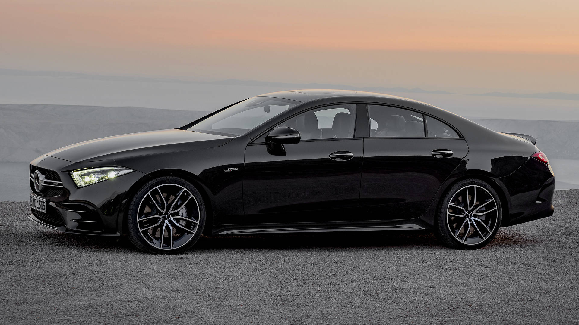 Mercedes Amg 桌布 壁紙 — 一輛時尚的黑色 Mercedes-AMG CLS 轎車在柔和的橙色日落天空下，在碎石觀景台上閃閃發光。