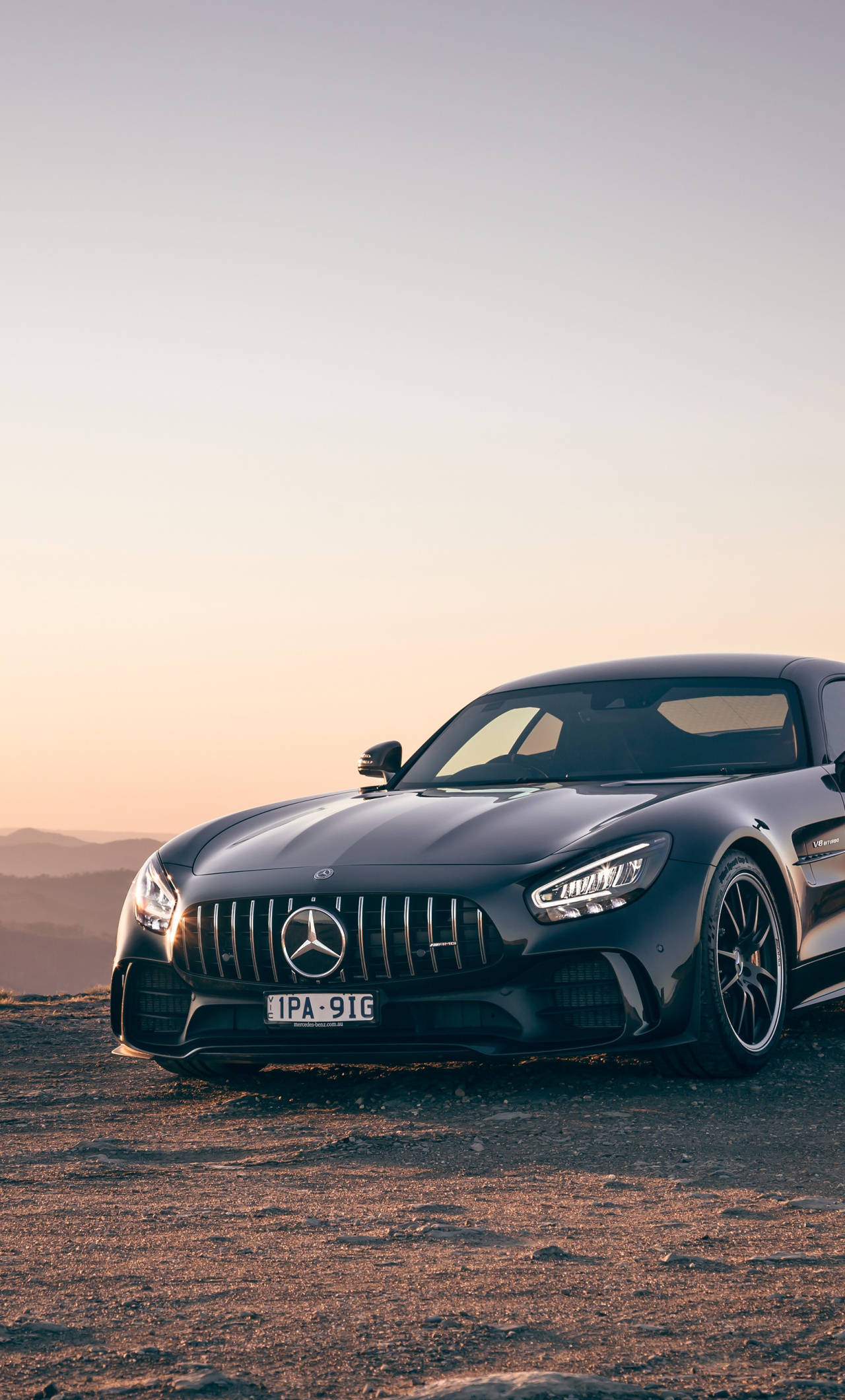 AMG iPhone 桌布 壁紙 — 一輛時尚的黑色 Mercedes-AMG GT 停在日落時分的塵土飛揚的山坡上，柔和的金色光線突出了其空氣動力學曲線和激進的格柵。