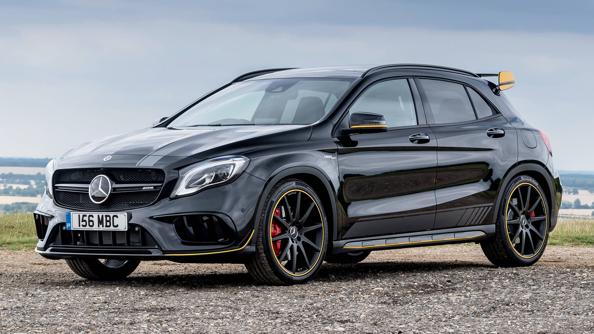 amg iPhone 桌布 壁紙 — 一輛時尚的黑色 Mercedes-AMG GLA45 S 停在礫石上，帶有黃色點綴和紅色制動卡鉗，天空多雲。