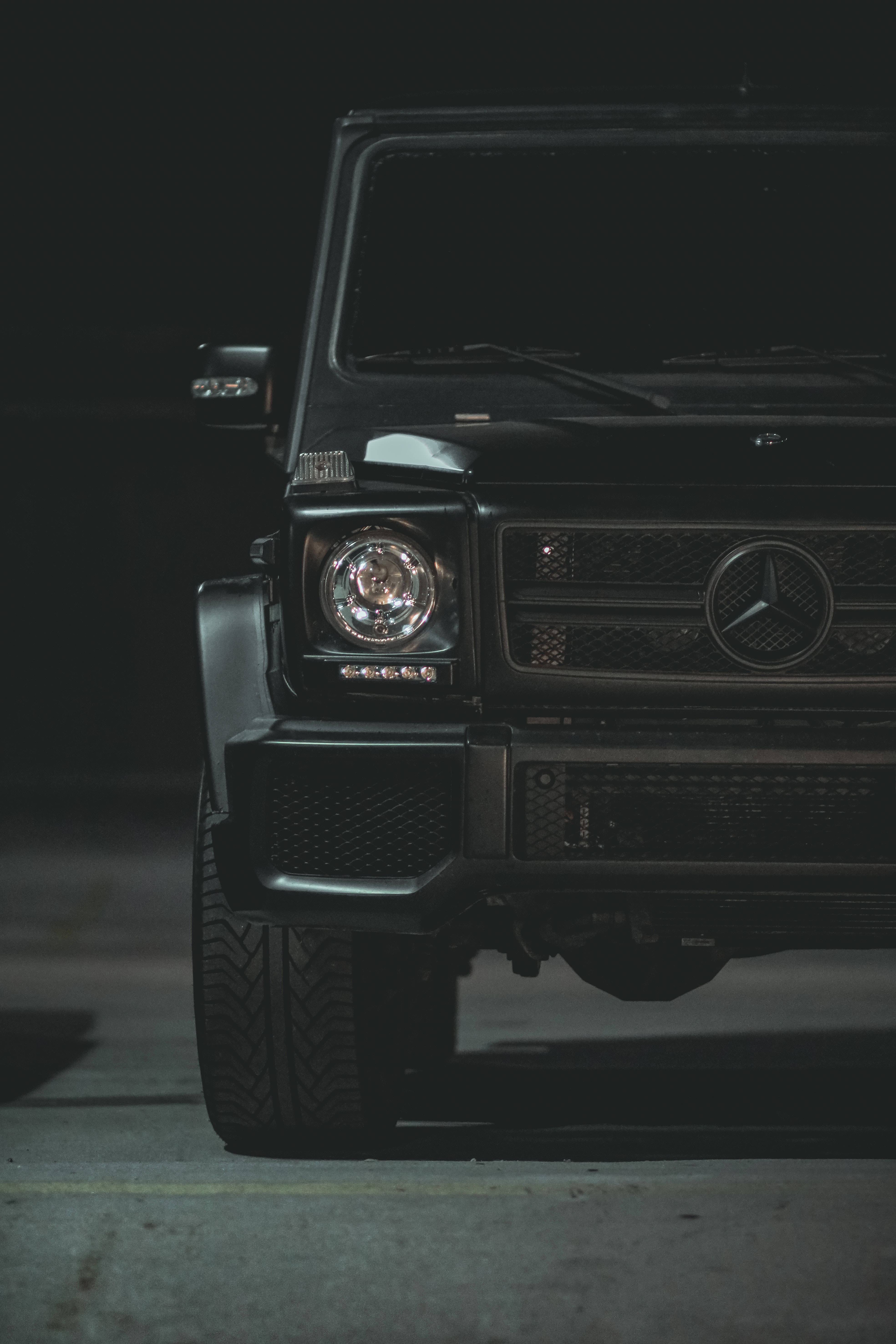 mercedes amg iPhone 桌布 壁紙 — 一輛時尚的黑色 Mercedes G-Wagen 在昏暗燈光下閃耀，車頭燈亮起，以黑暗的城市背景為襯托。