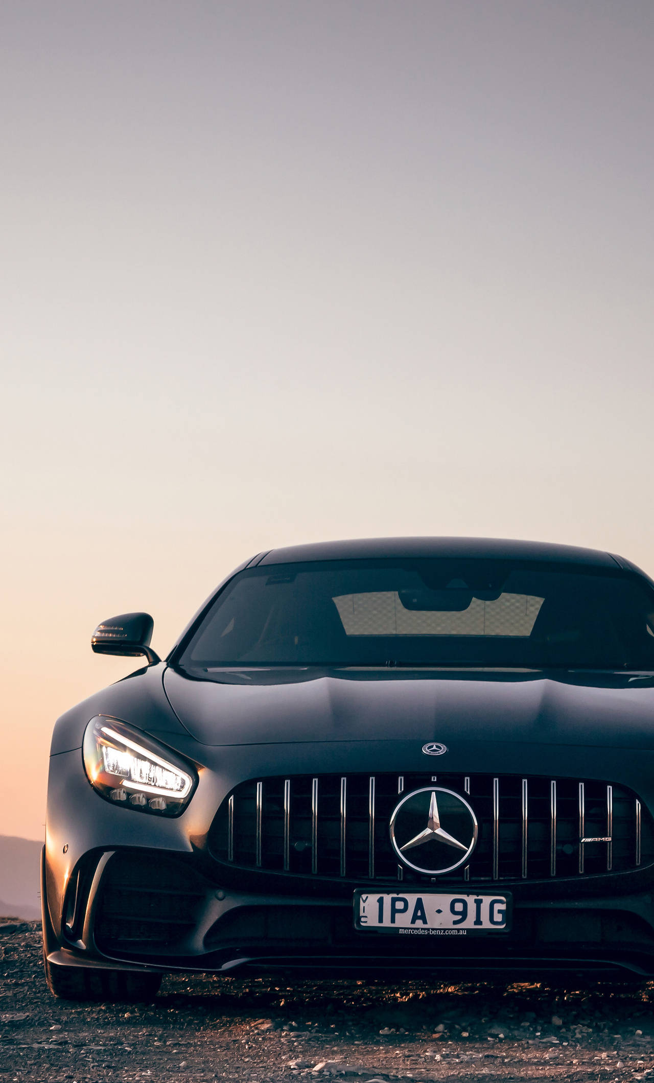 amg iPhone 桌布 壁紙 — 一輛時尚的黑色 Mercedes-AMG GT 停在夕陽下的礫石上，溫暖的環境光凸顯其激進的前格柵和鋒利的大燈。