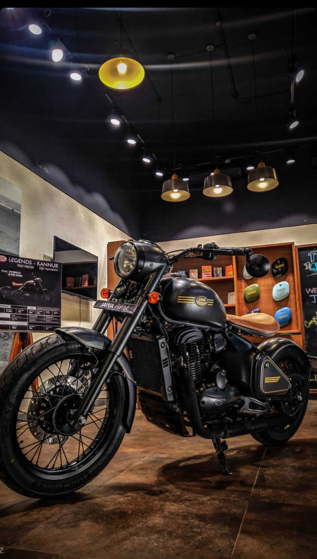 Bobber 桌布 壁紙 — 一輛時尚的黑色 Royal Enfield 摩托車在溫暖的吊燈下閃閃發光，位於一個配有木架和復古裝飾的時尚室內展廳。