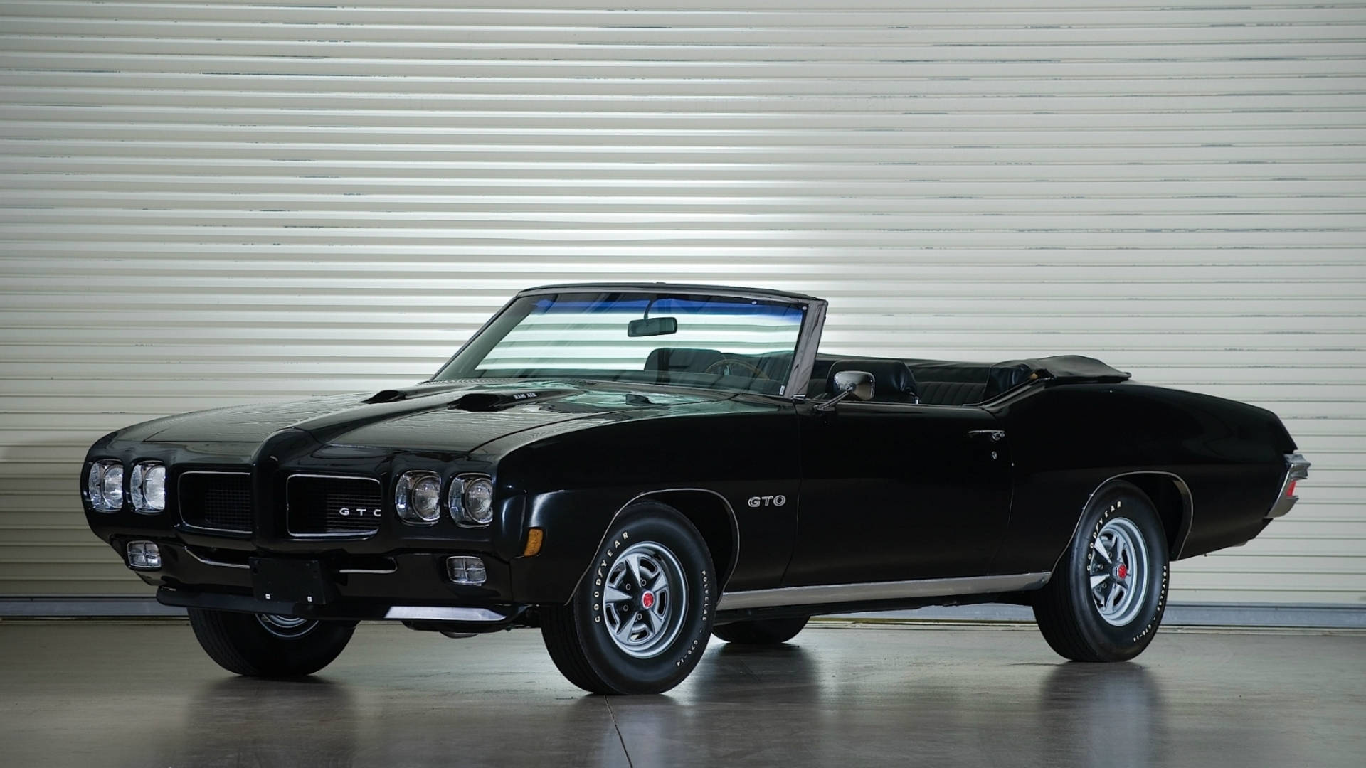 Pontiac 桌布 壁紙 — 一輛時尚的黑色1970年代龐蒂亞克GTO敞篷車，配備鍍鉻輪圈和軟頂，停放在白色波紋金屬車庫門前。