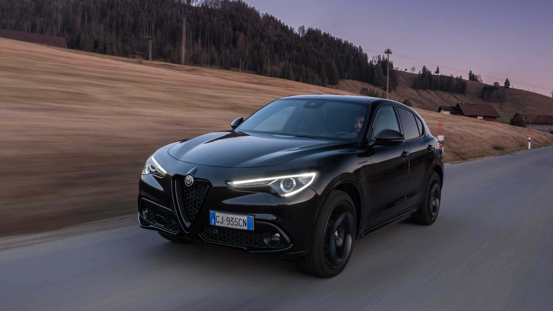 愛快羅密歐 桌布 壁紙 — 一輛時尚的黑色Alfa Romeo SUV在黃昏時分沿著蜿蜒的鄉村道路疾馳，背景是模糊的金色田野和森林覆蓋的山丘。