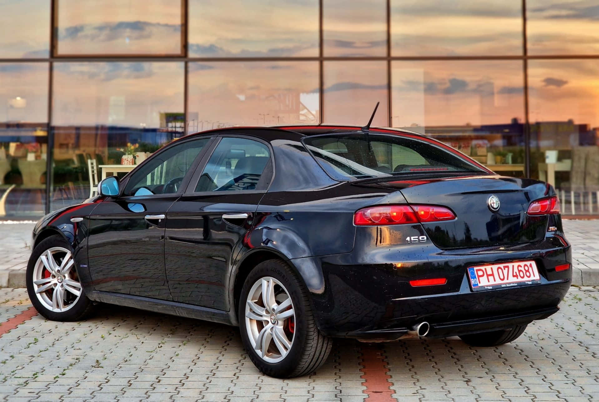 Alfa Romeo 159 桌布 壁紙 — 一輛時尚的黑色Alfa Romeo 156轎車停放在鋪砌的廣場上，夕陽西下，其光澤表面反射著溫暖的橙色天空。